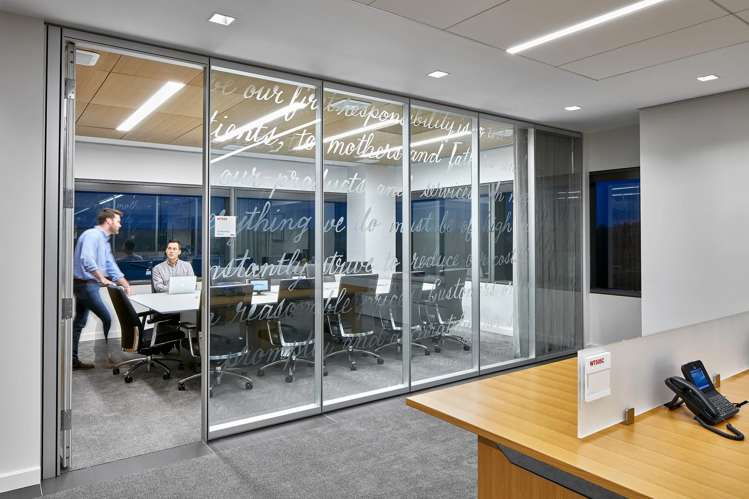 Office conference room with glass walls featuring cursive writing, two men engaged in conversation, one seated with a laptop and the other walking, modern furnishings, white walls, ceiling lights, and wood accents.