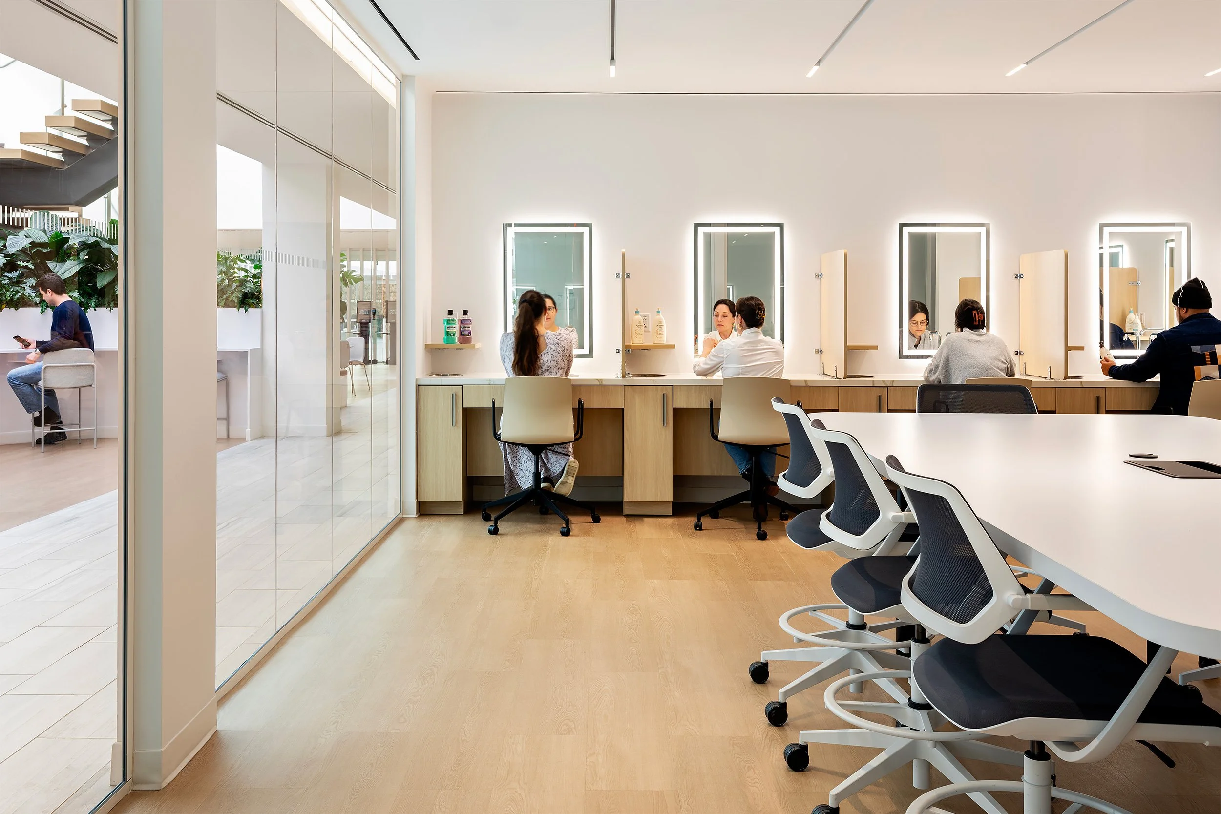 A modern salon with multiple styling stations, illuminated mirrors, and several people getting their hair or makeup done.