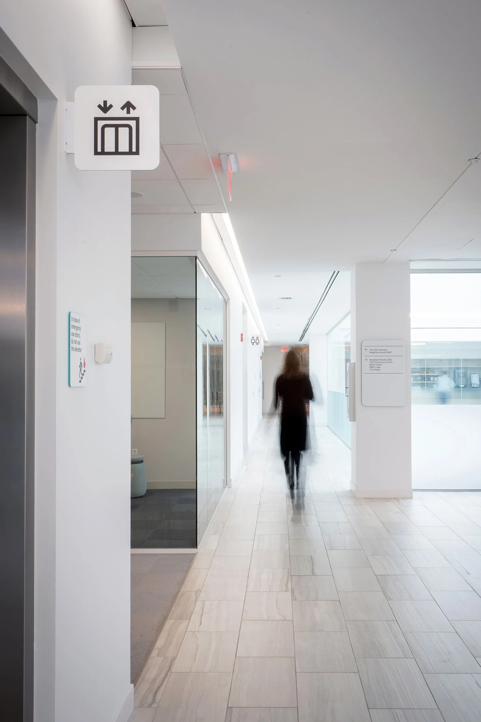 A woman walking down a bright, modern indoor corridor with clean white walls and light-colored tiled floor, near an elevator with an upward and downward arrow sign.