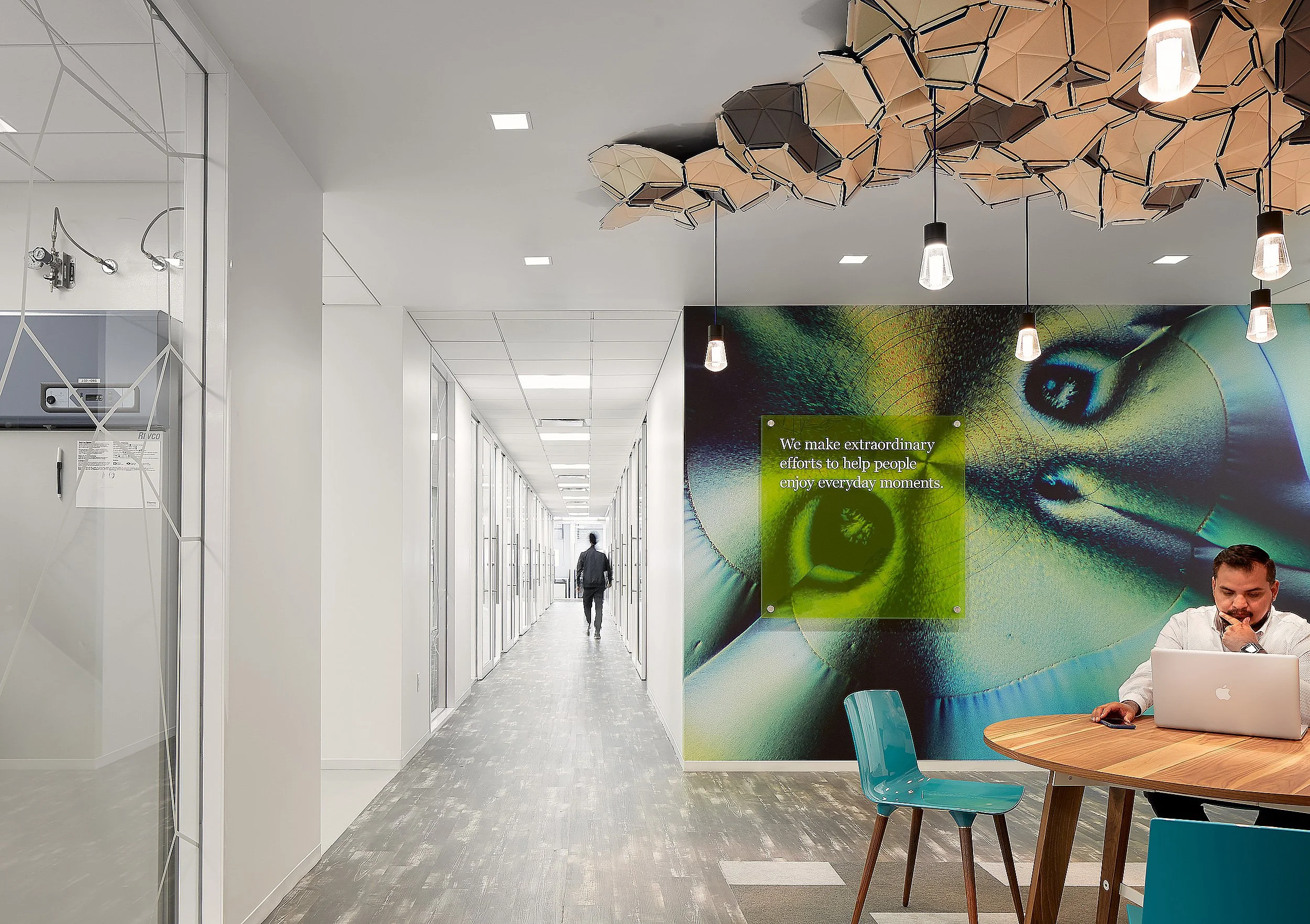 Modern office interior with white walls, a long corridor, and a lounge area featuring a large wall mural of a peacock's eye and a green sign stating, "We make extraordinary efforts to help people enjoy everyday moments." There are pendant lights hanging from a geometric ceiling installation, a woman walking down the corridor, and a person sitting at a table using a laptop.