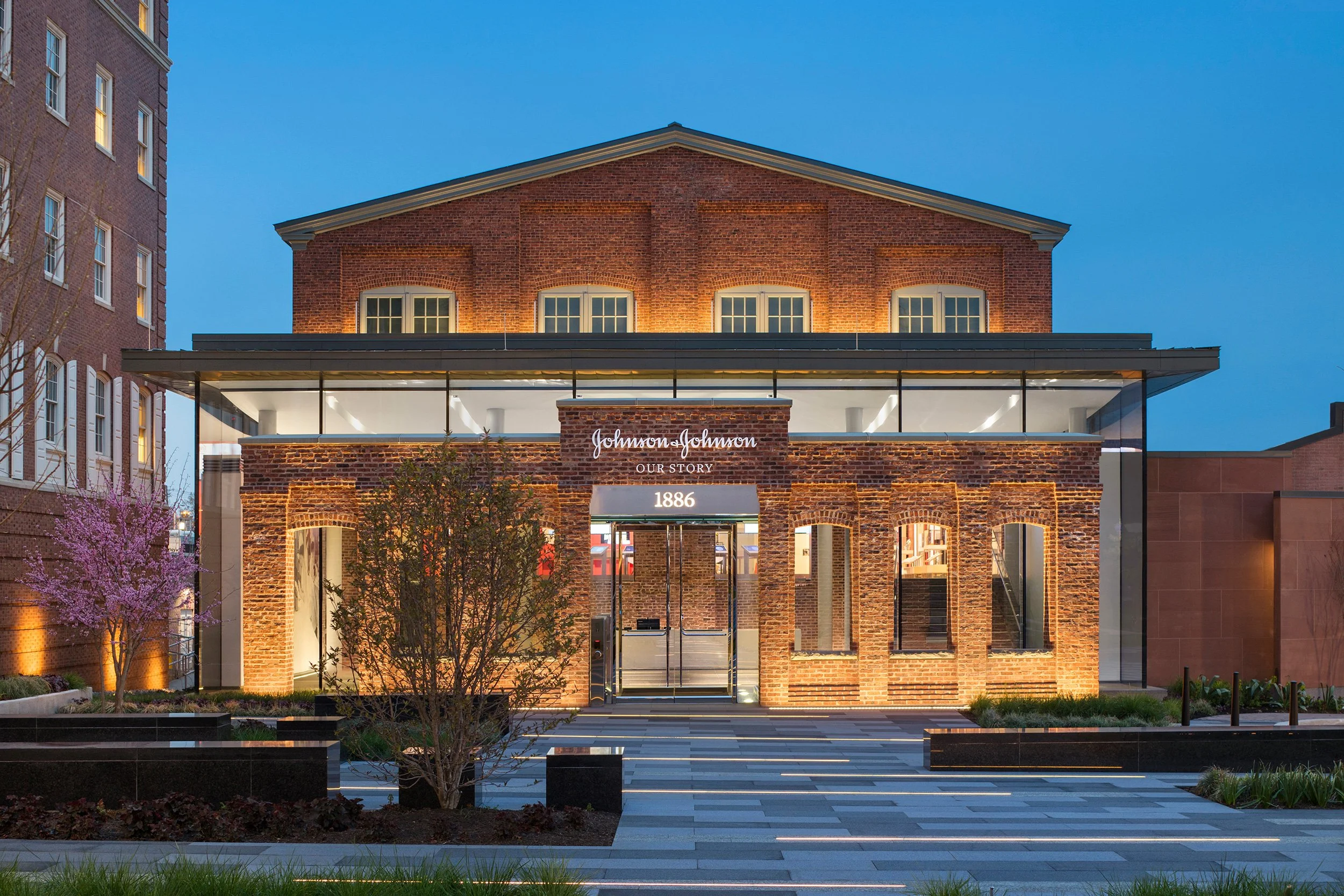 Facade of Johnson & Johnson building with brick walls, illuminated windows, glass entrance, and a sign with the company's name and founding year 1886. Evening setting with lit trees and plants.