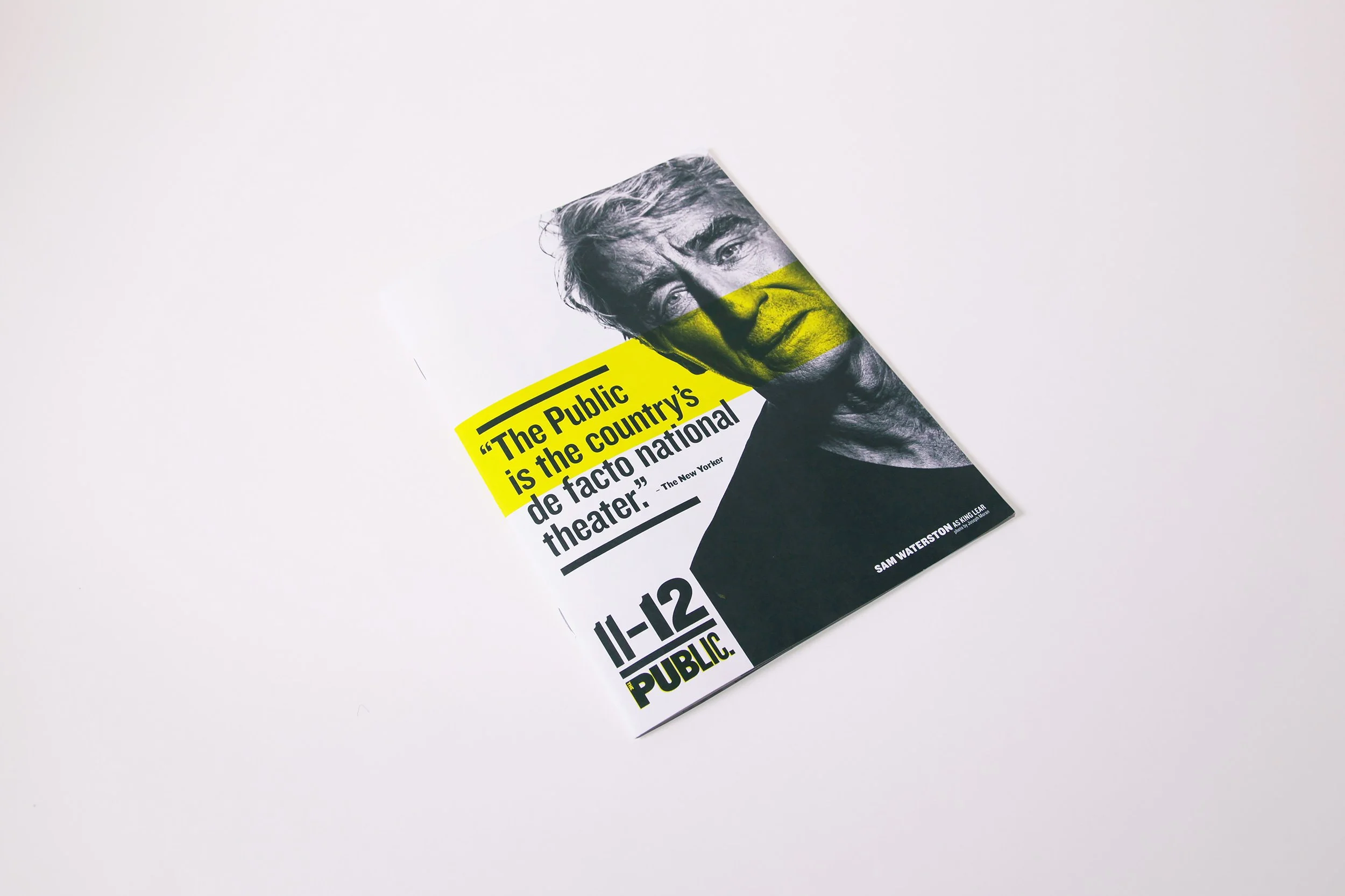 Close-up of a magazine or brochure featuring a black and white photo of a man's face with a yellow strip across it. The text on the yellow strip reads, "The Public is the country's de facto national theater." The magazine is placed on a plain white background.