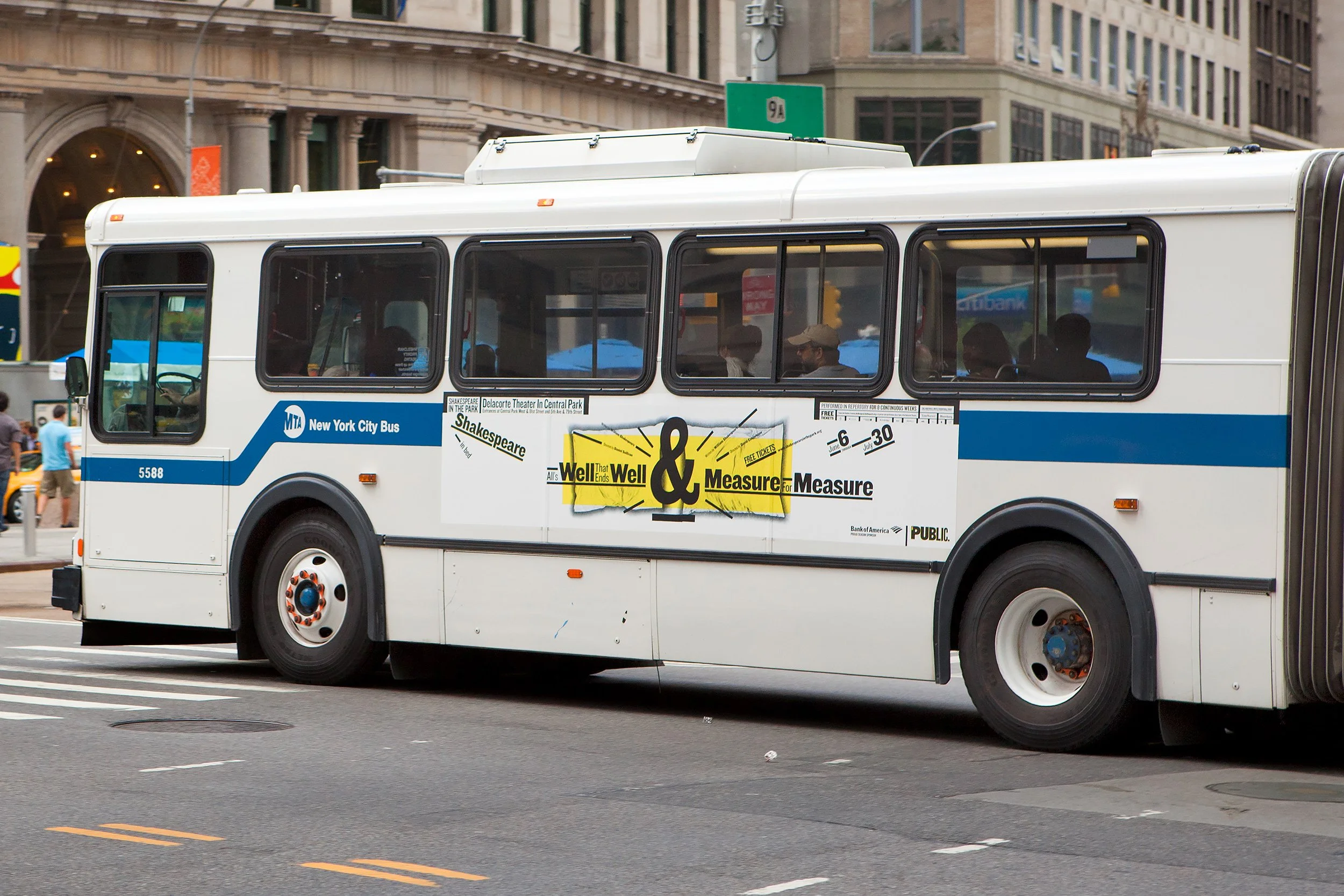 A white New York City MTA bus with an advertisement for a Shakespeare play on the side, driving on a city street with buildings and pedestrians visible in the background.
