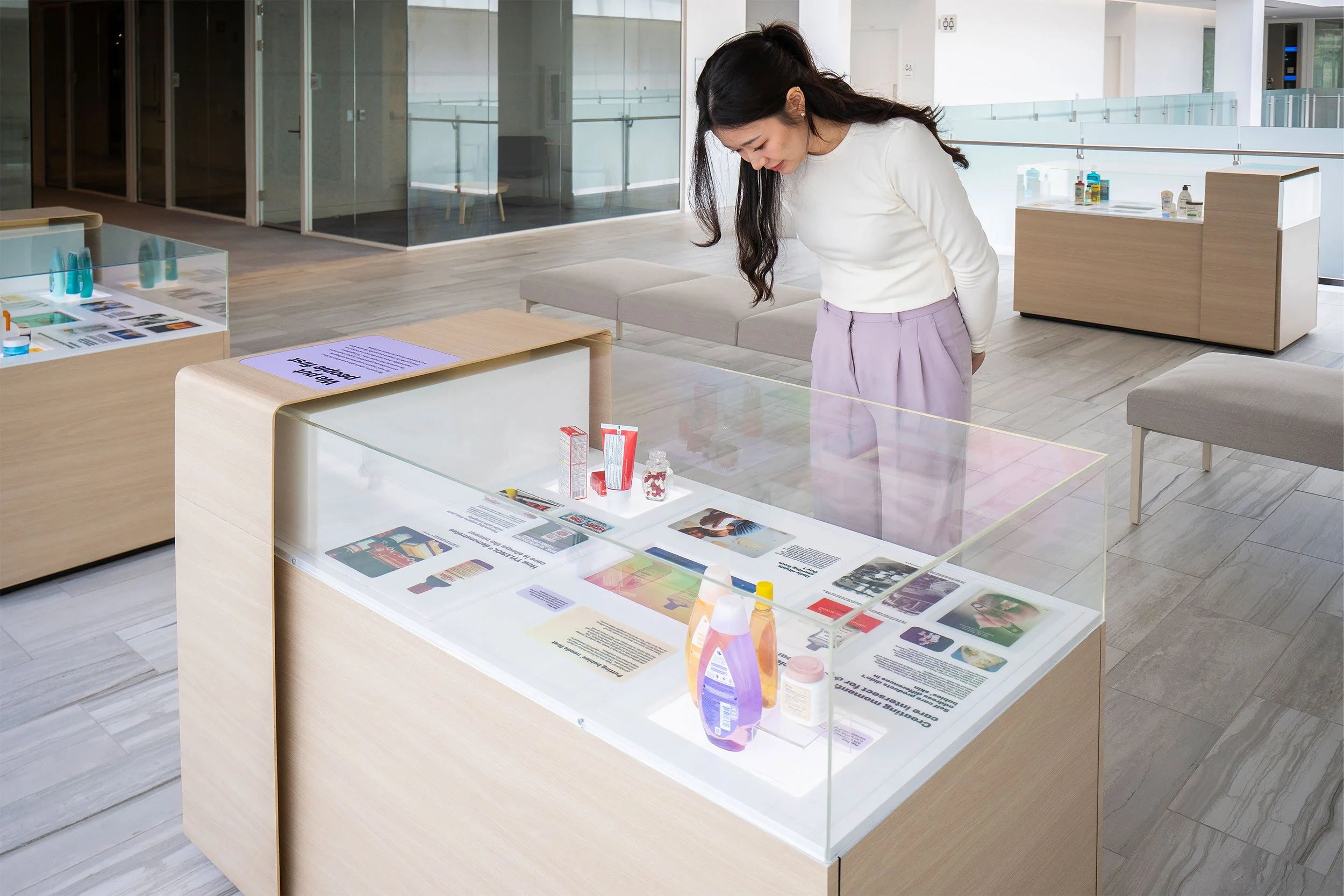 A woman in a white long sleeve top and purple pants is looking down and examining items inside a glass display case in a modern, bright interior space with wooden furniture and glass walls.
