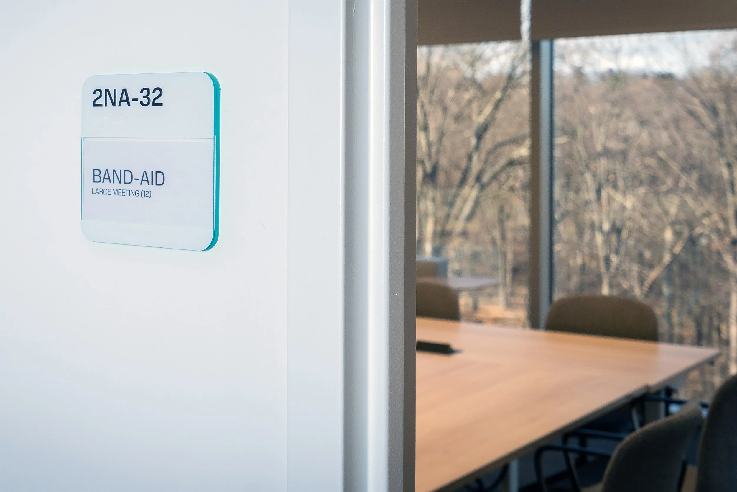 Close-up of a sign on a wall reading '2NA-32 BAND-AID LARGE MEETING (12)', with a room visible in the background through a window showing chairs and trees outside.
