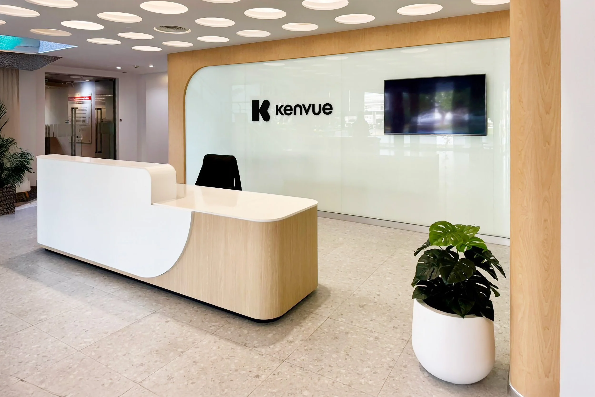 Modern office lobby with a reception desk, a large wall-mounted TV, and potted plants. The wall has a logo that says 'Kenvue'.