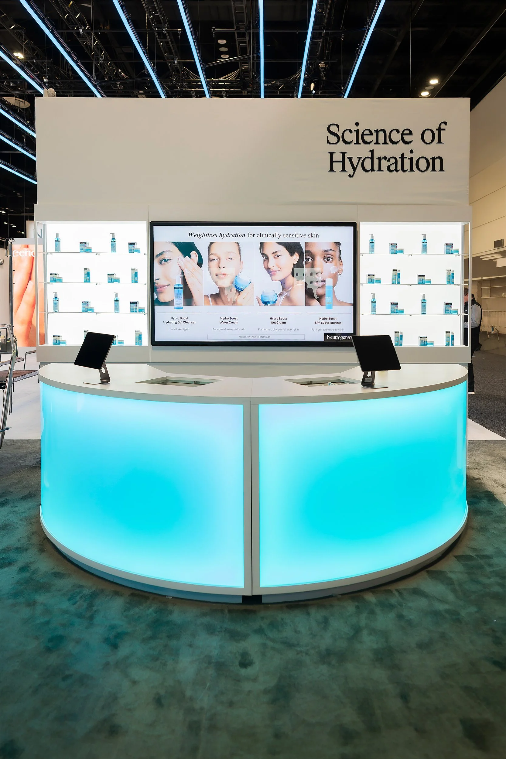 A booth titled 'Science of Hydration' showcasing Neutrogena skincare products with product displays on illuminated shelves and a digital screen featuring models demonstrating hydration routines.