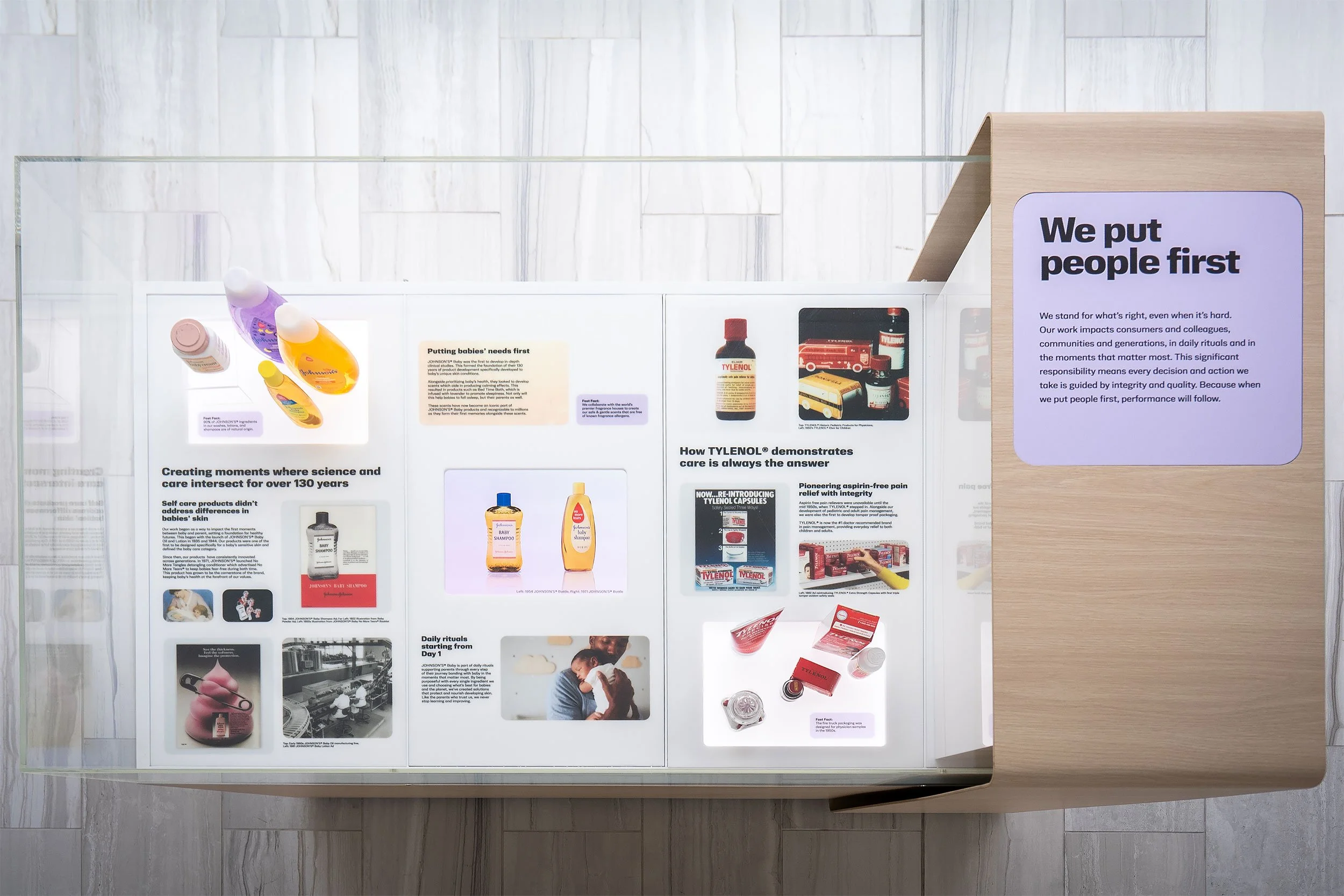 Display board featuring various pharmaceutical and healthcare product advertisements, including bottles, boxes, and informational text, encased in a glass frame on a light-colored wooden stand.