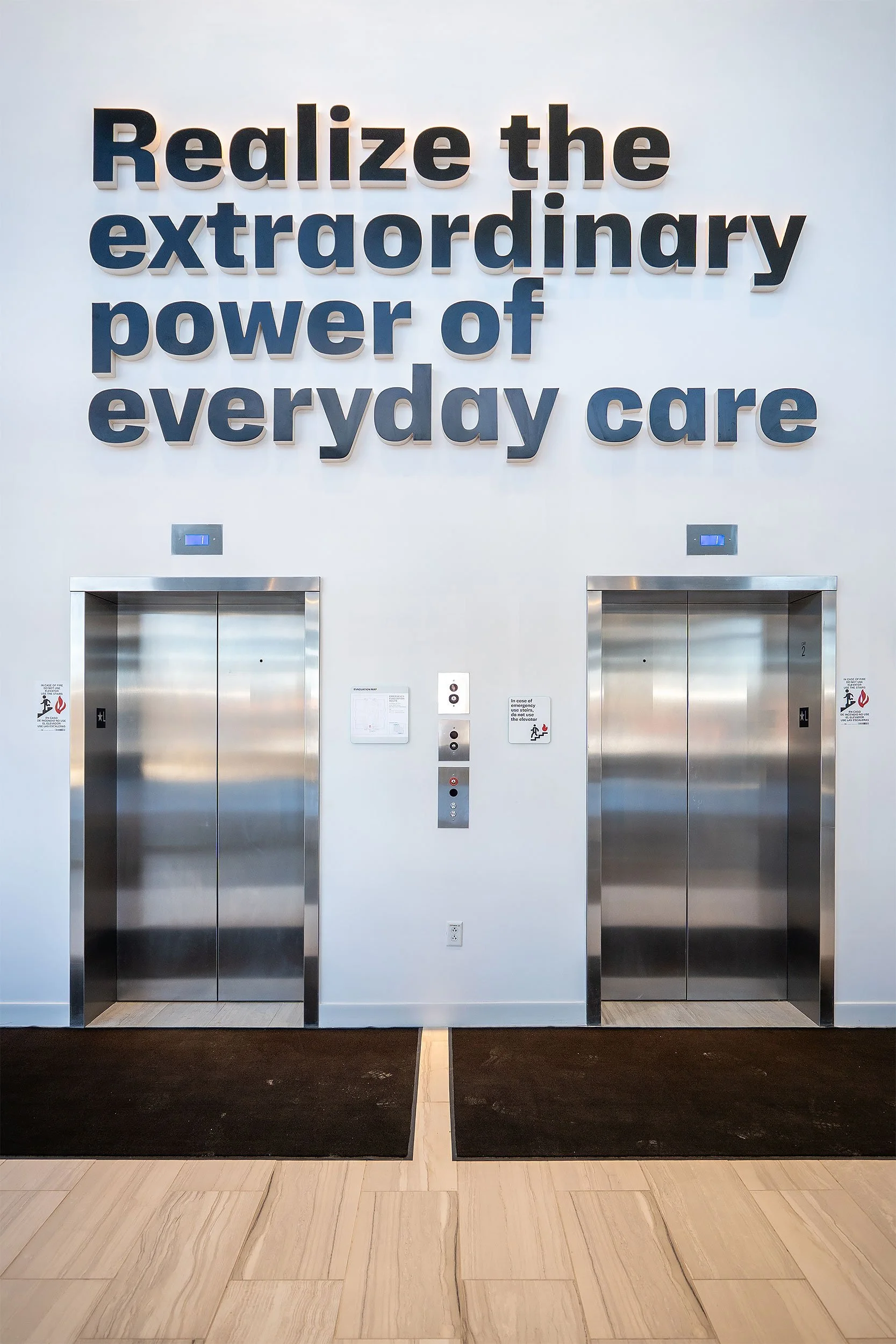 A wall with large black 3D letters reading, 'Realize the extraordinary power of everyday care,' with two elevator doors below and control panels and signage near the doors.