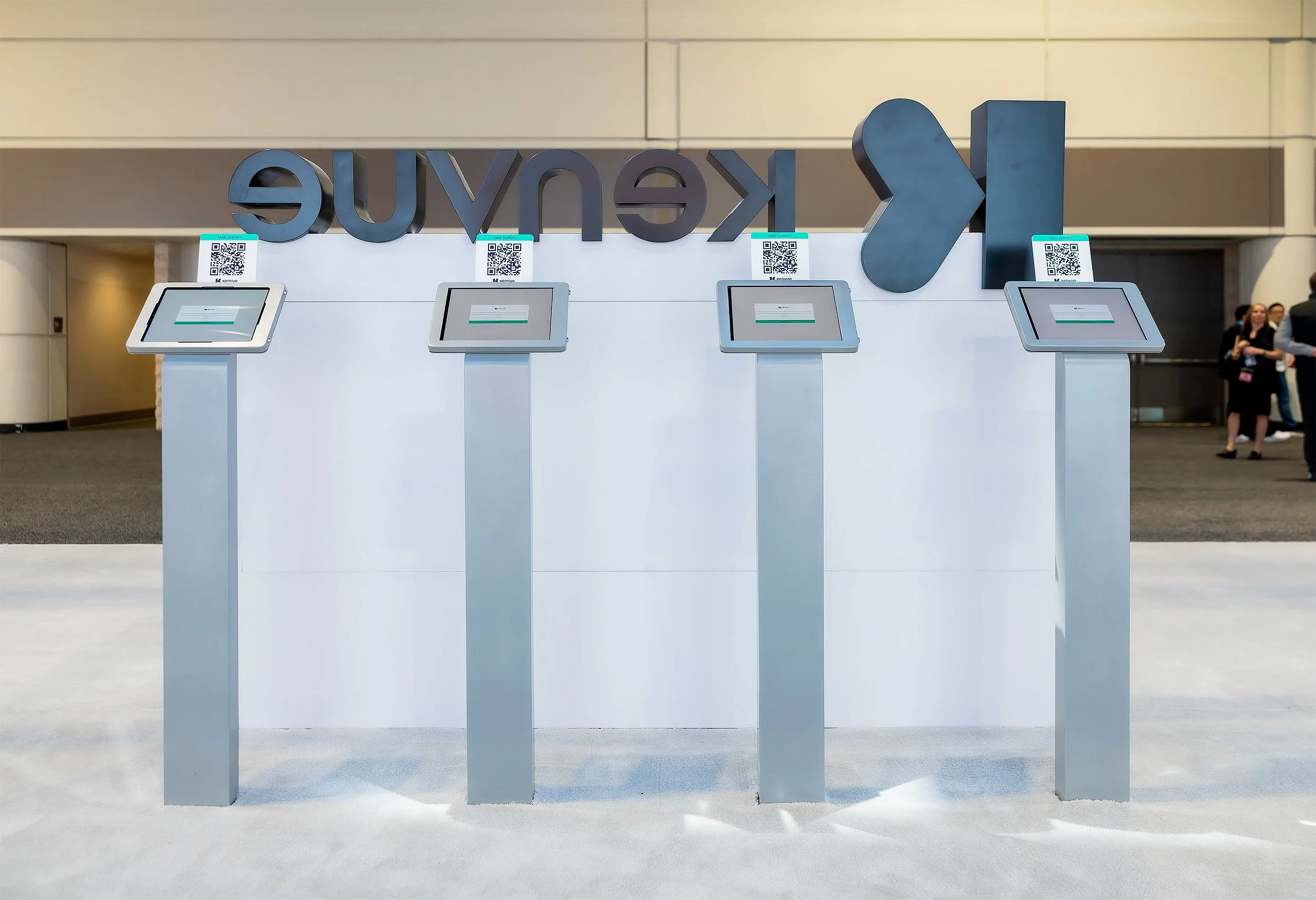 Photo of four white kiosk booths with touchscreen monitors and QR codes, in front of a large black sign with stylized letters and arrows, at an indoor queue area.