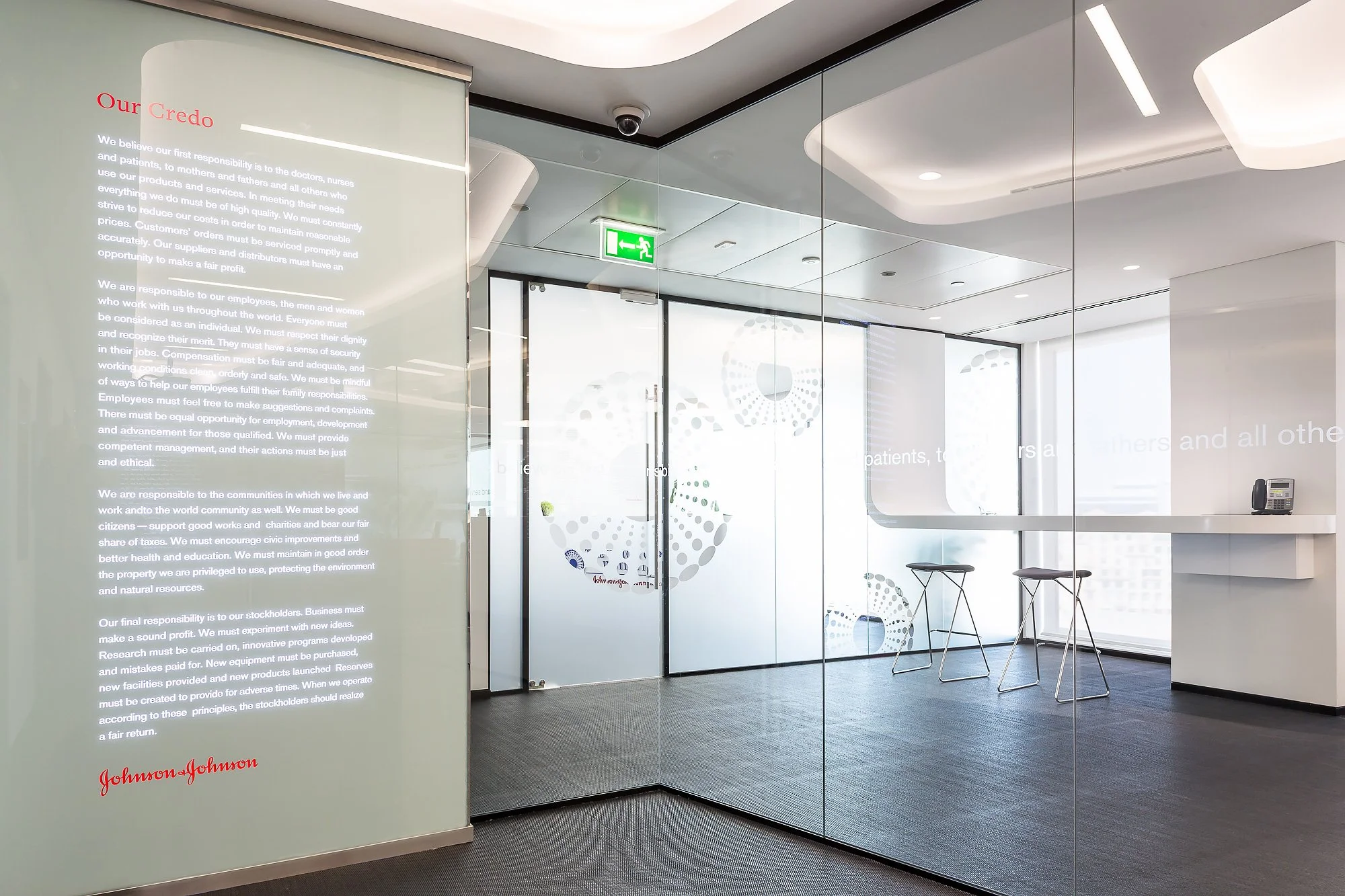 A modern office space featuring glass walls, a digital display, and a seating area with stools. There is an emergency exit sign on the ceiling and a control telephone on a small table.