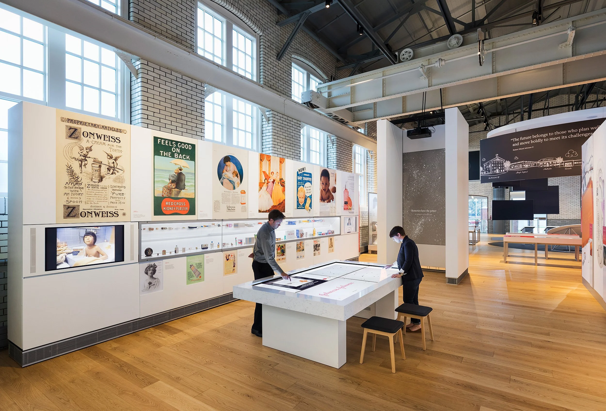 Interior of a museum or exhibition space with large windows, displaying vintage advertisements and informational displays, where two people are examining exhibits at a white table.