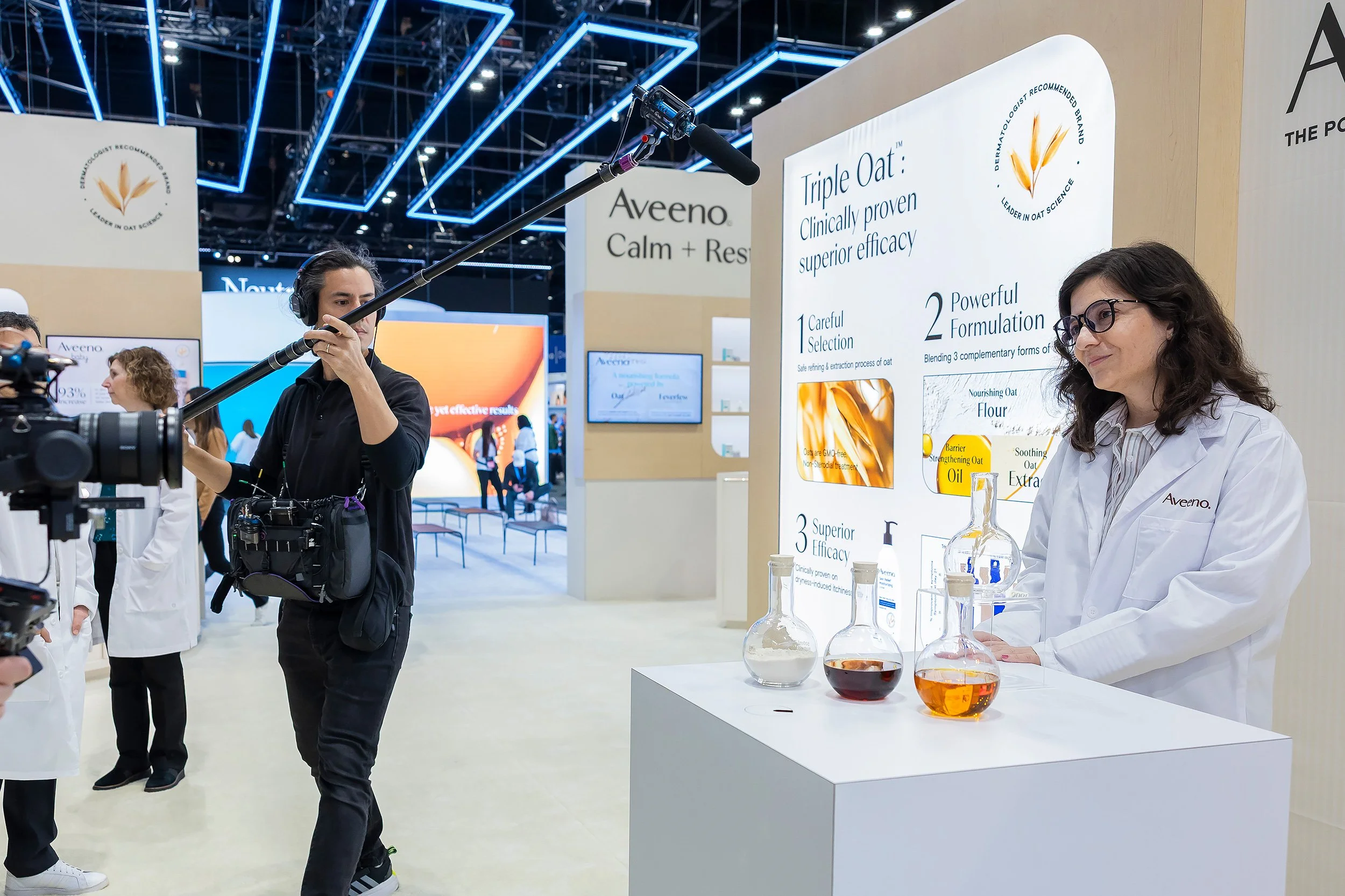 A woman in a white lab coat demonstrating skincare products at a trade show booth, with informational posters about oat ingredients and efficacy, and a person holding a boom microphone recording the event.