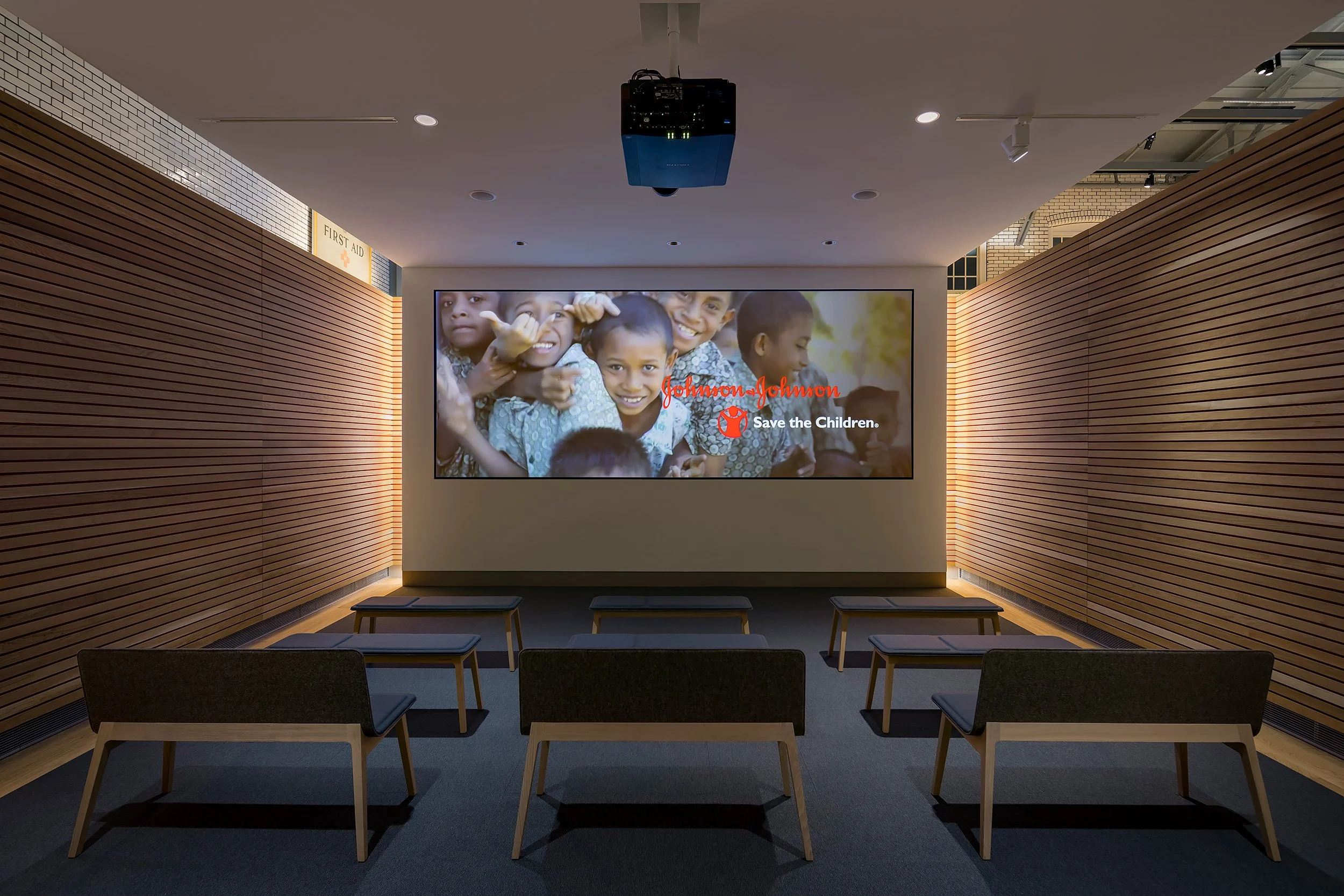 Empty presentation room with wooden walls, a large screen displaying children and the Johnson & Johnson logo for a charity campaign, and empty chairs.