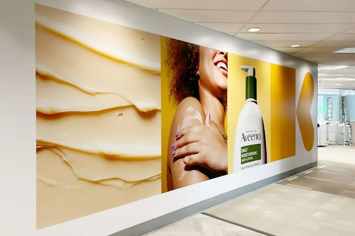 A large advertisement mural on a wall inside a building, showing a woman with curly hair smiling, hugging herself, and a bottle of AloeNo aloe vera product on a yellow background.