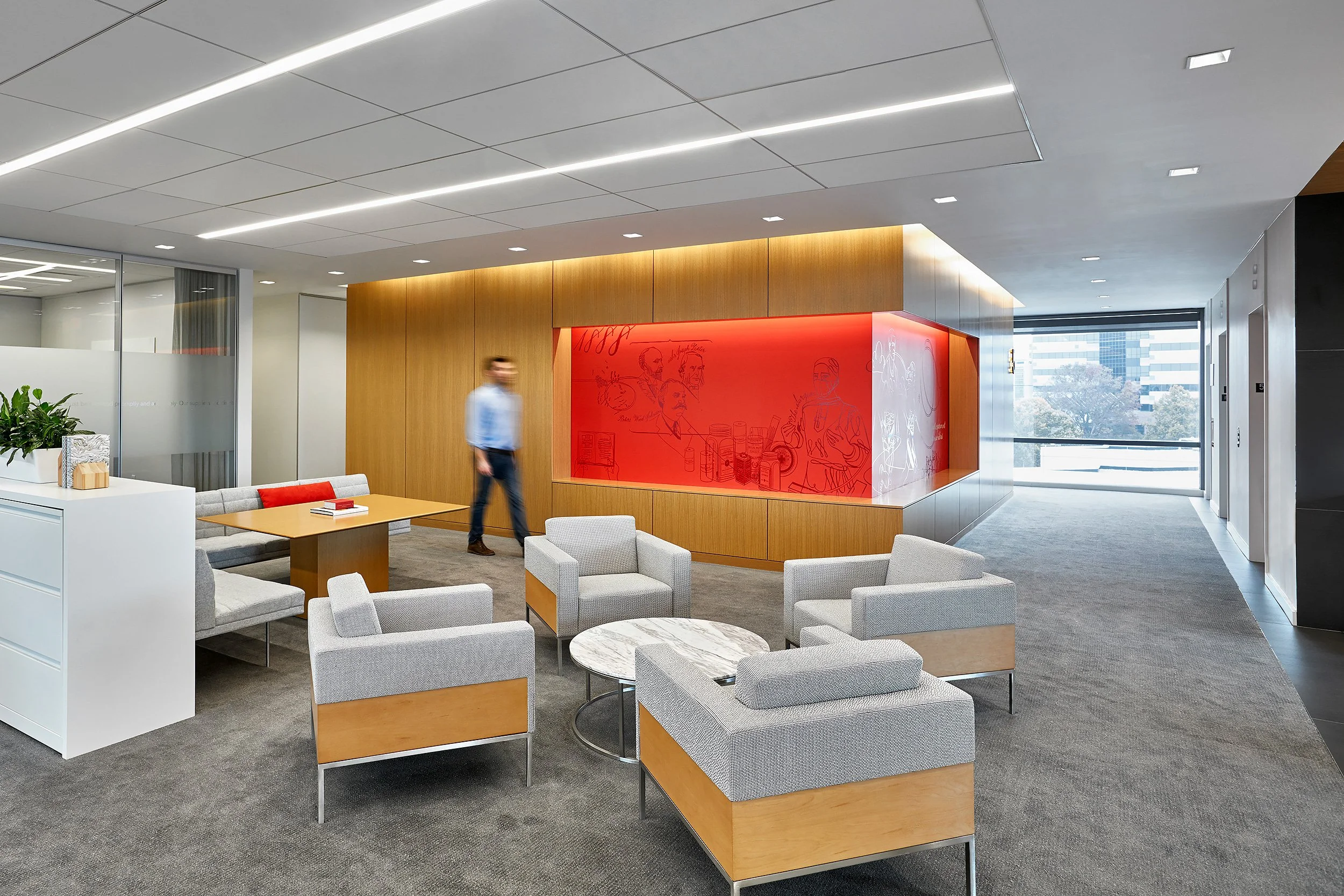 Modern office lounge area with gray upholstered chairs, a wooden table, a gray sofa with red accent pillows, a white side table, and a red wall display with sketches of people and objects, large windows showing city buildings outside.