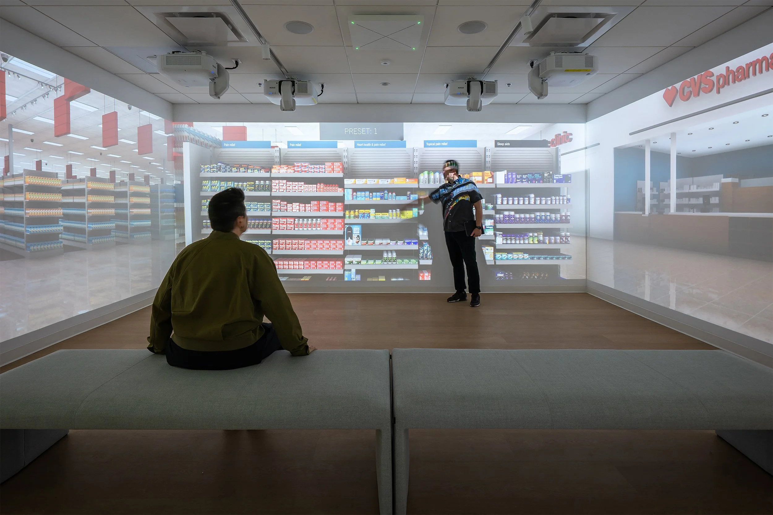 Inside a pharmacy with two people, one sitting on a bench and one standing pointing at a display shelf of medicine.