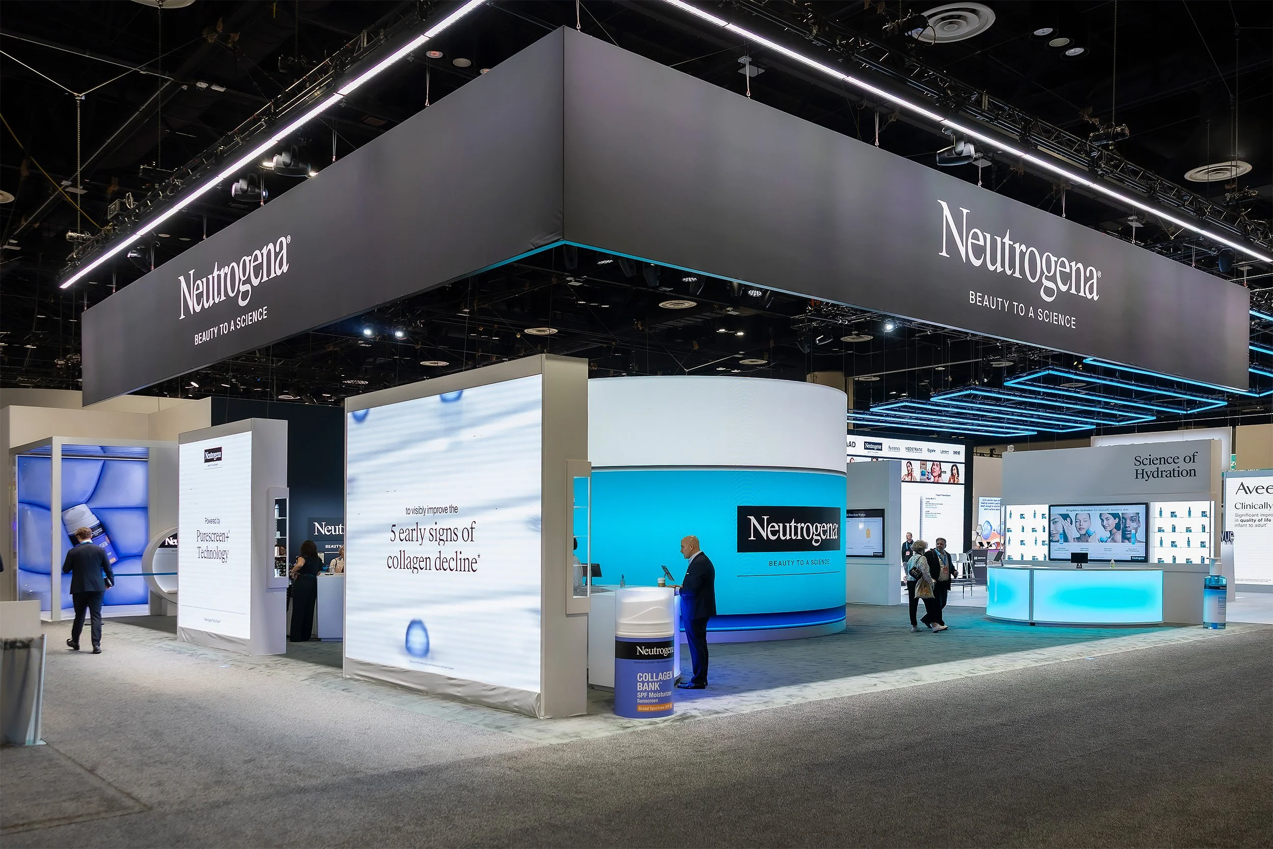 Trade show booth for Neutrogena with multiple display screens and informational signage, visitors walking around, in a large convention center setting.