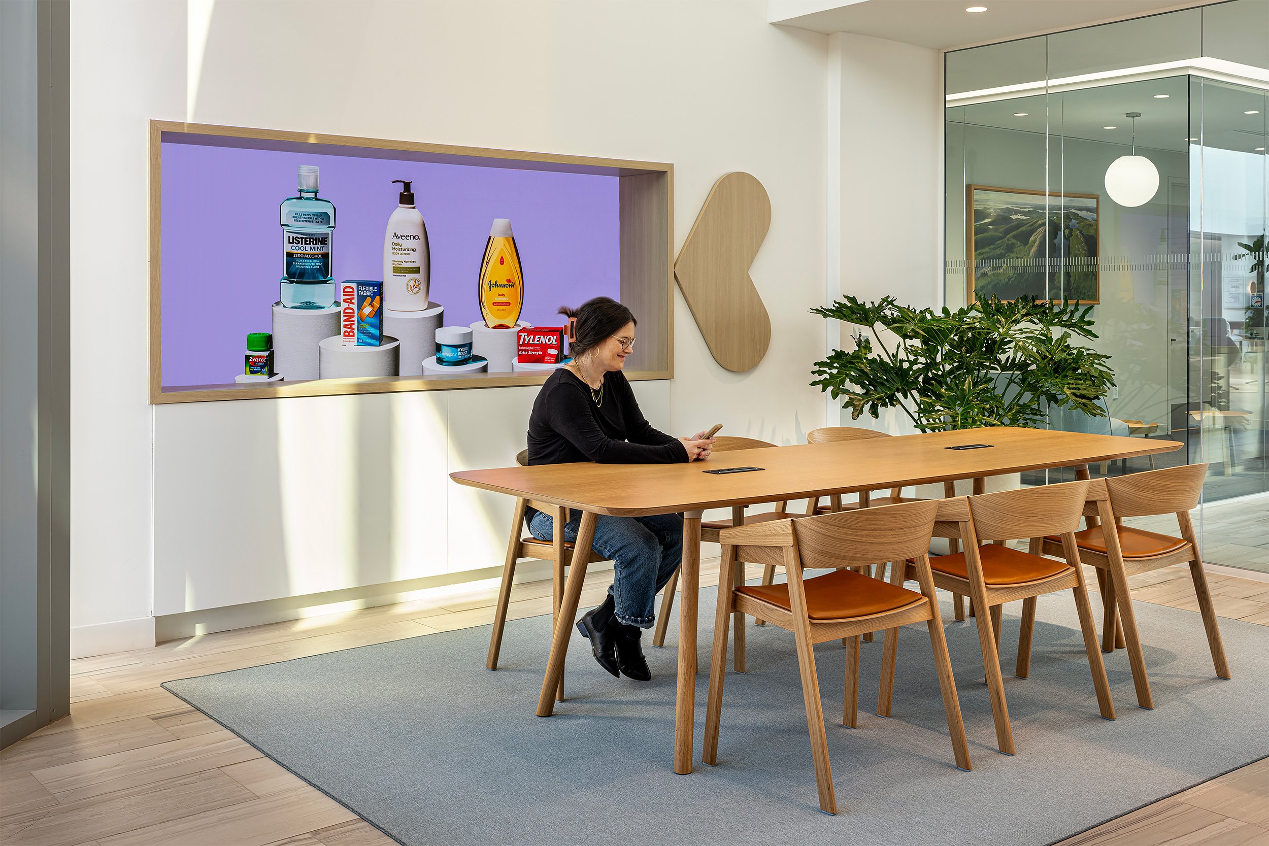 A woman sitting at a long wooden table, looking at her phone in a modern, well-lit conference or meeting room with a large plant and glass wall.