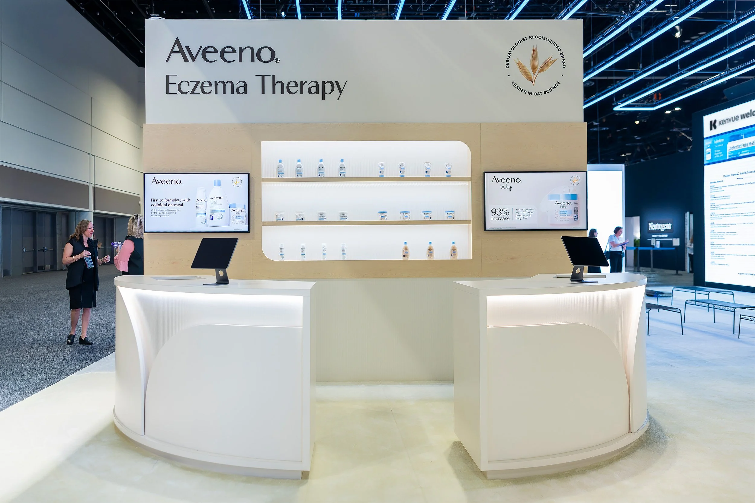 A booth for Aveeno eczema therapy at a trade show or conference. The booth has a large sign with the Aveeno logo and the words "Eczema Therapy." There are digital screens displaying product information on either side of a display of Aveeno products, with shelves backlit with white light. Two women are standing near the booth, one holding a water bottle and the other engaged in conversation in the background.