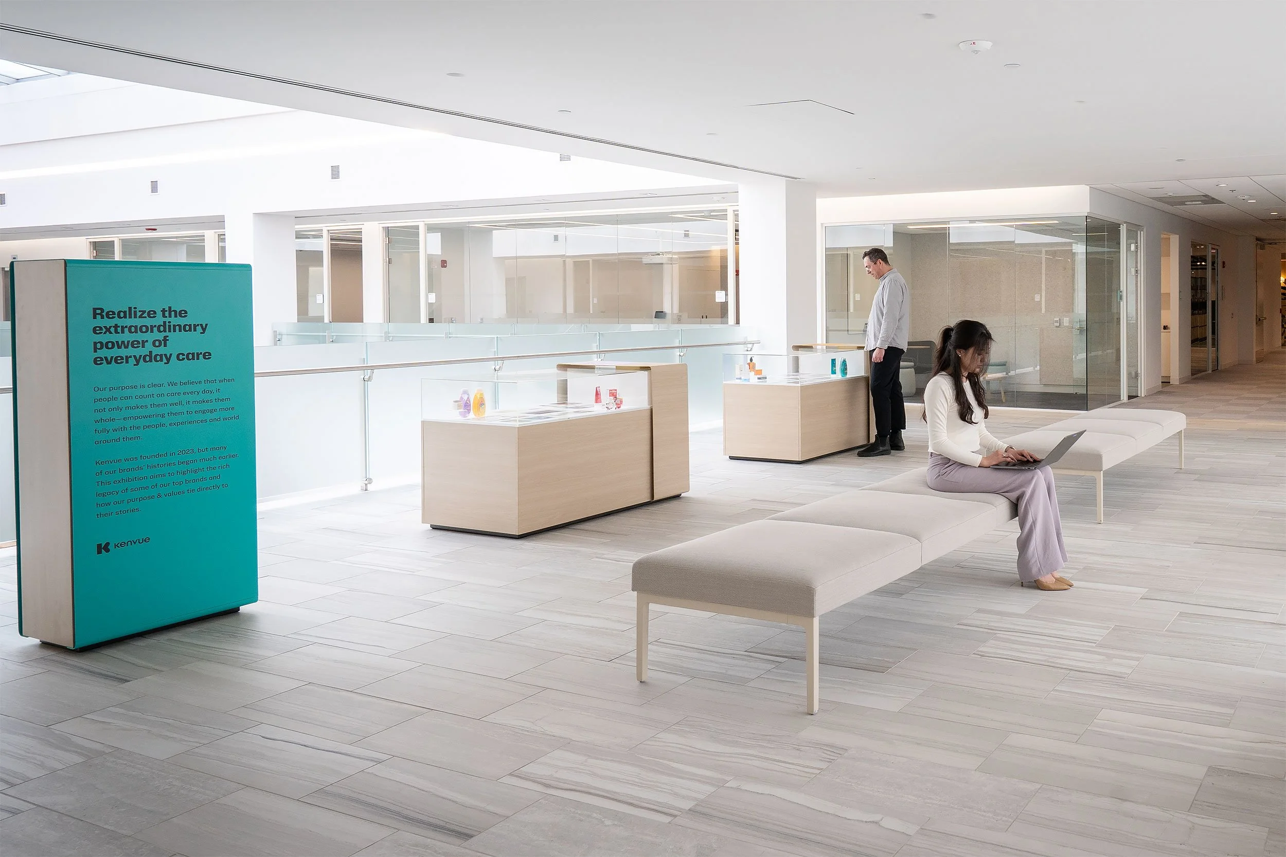 A modern, minimalistic lobby or waiting area in a corporate or healthcare building. There is a teal informational sign and two people: a man standing and a woman sitting on a bench using a laptop, with glass walls and display cases of products.