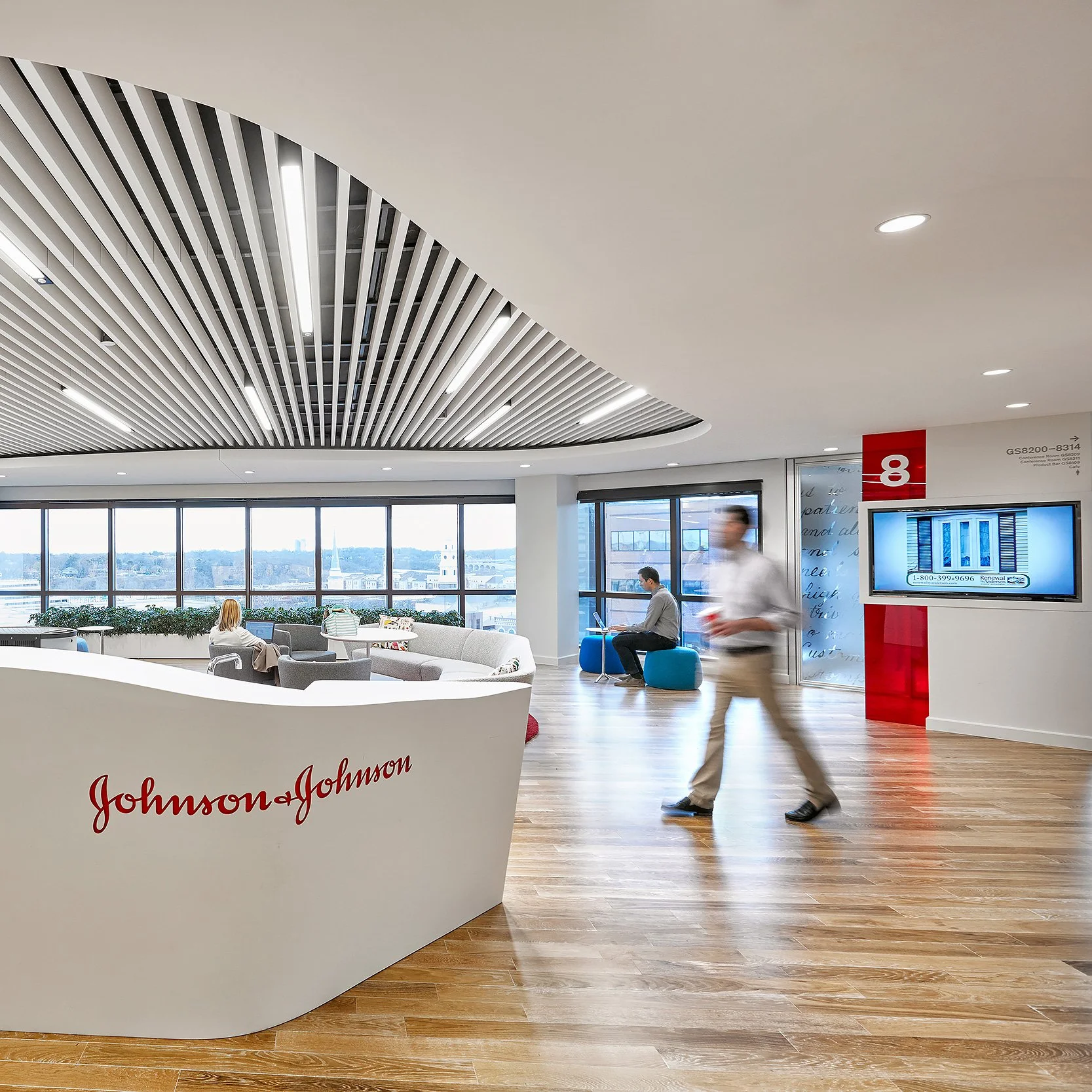 A modern office lobby with the Johnson & Johnson logo on a white curved reception desk, wooden flooring, large windows with a city view, and people seated and walking in the space.