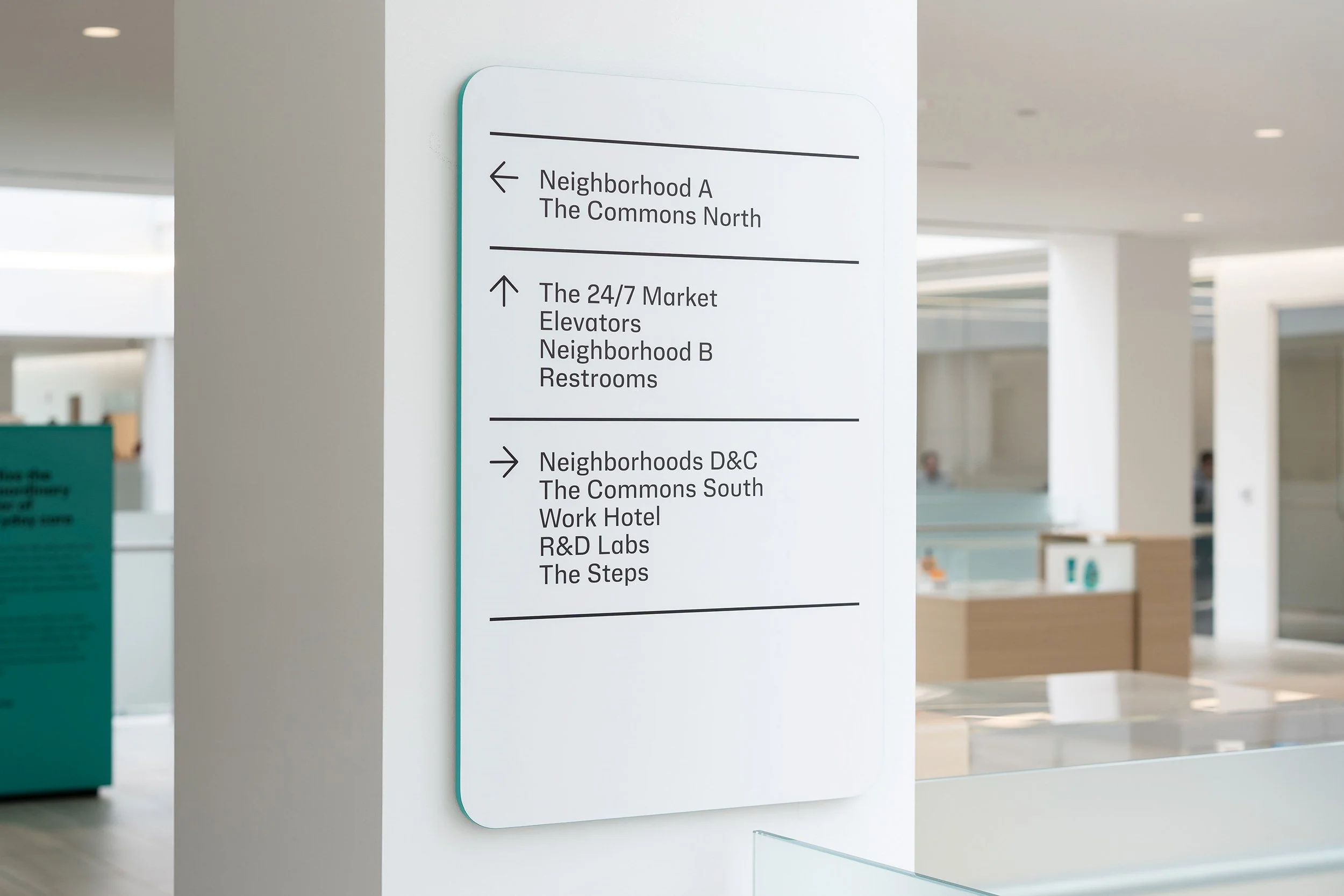 Interior of a building with a white directory sign indicating directions to various neighborhoods and facilities, including Neighborhood A, The Commons North, The 24/7 Market, Elevators, Neighborhood B, Restrooms, D&C, The Commons South, Work Hotel, R&D Labs, and The Steps.