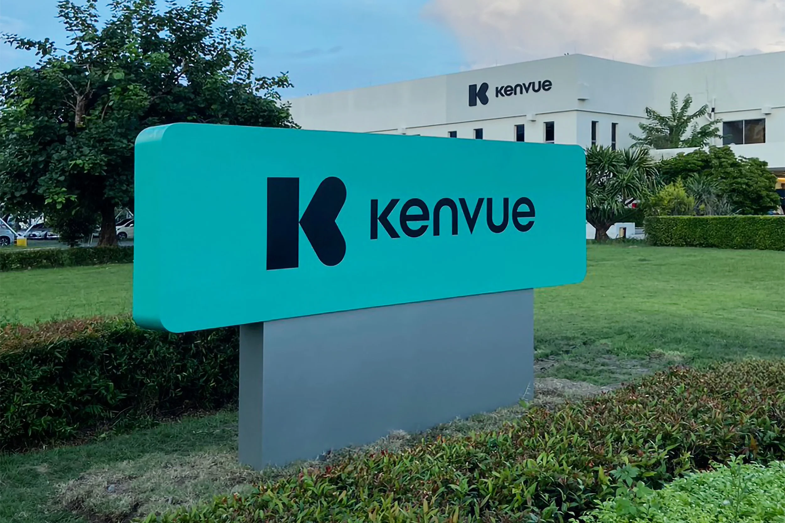 A large turquoise sign with the black logo and text 'Kenvue' is in the foreground of a landscaped area with grass and bushes. In the background, there is a white building with the 'Kenvue' logo on it, surrounded by trees and parked cars under a partly cloudy sky.