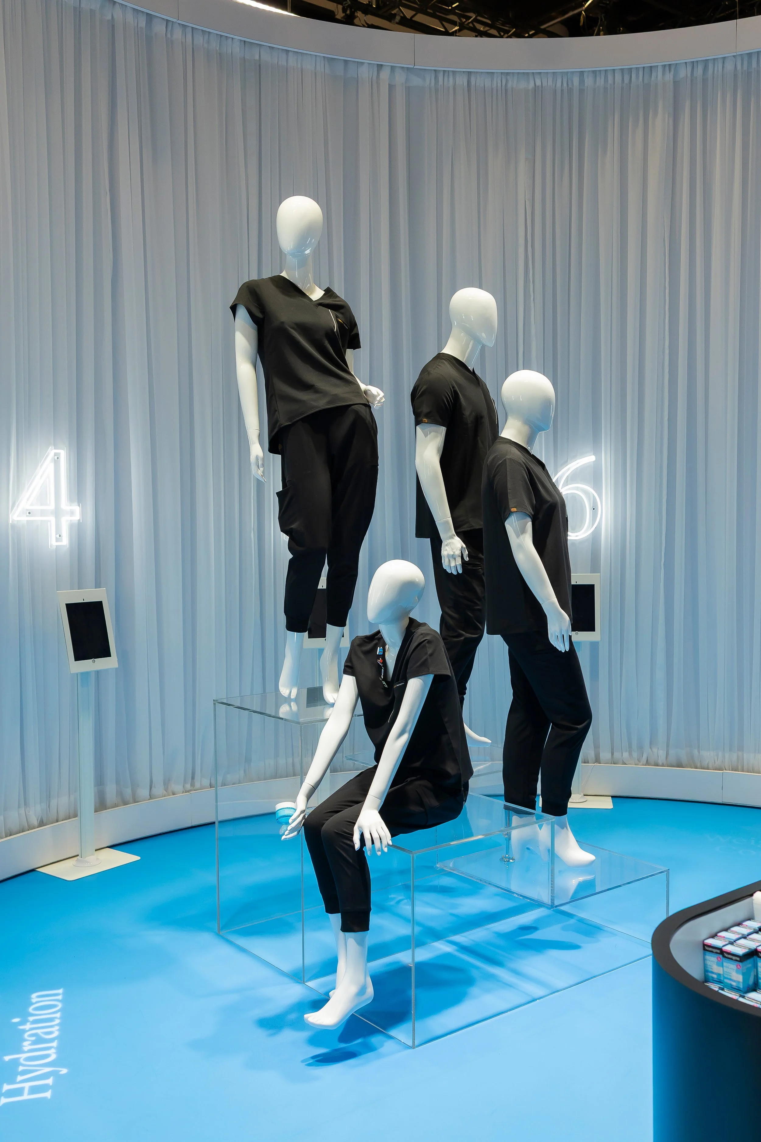 Display of mannequins dressed in black clothing, positioned on clear platforms, set against a backdrop of light blue curtains and illuminated with bright lighting at a retail store or exhibition.