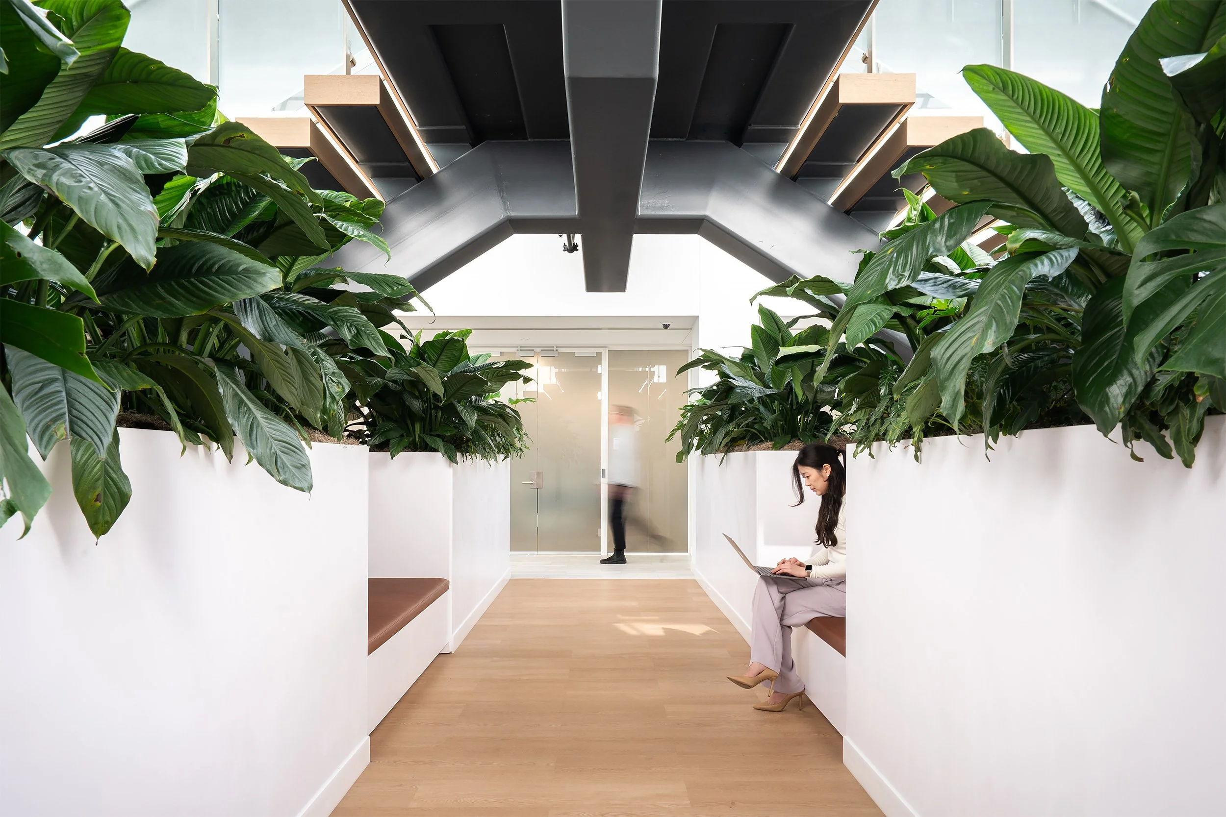 Indoor space with green plants on white walls, a woman in heels sitting on a bench working on a laptop, and a person walking in the background.