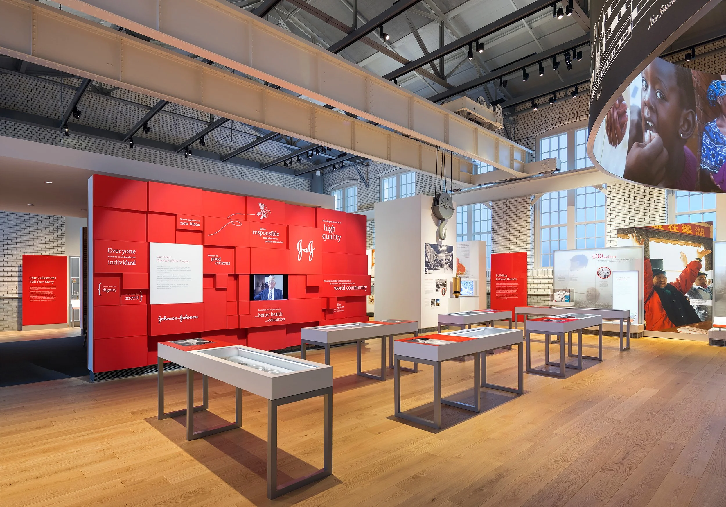 Interior of a museum or exhibition space with red display walls featuring educational information and images about Johnson & Johnson, with large windows, industrial ceiling, and display tables in foreground.