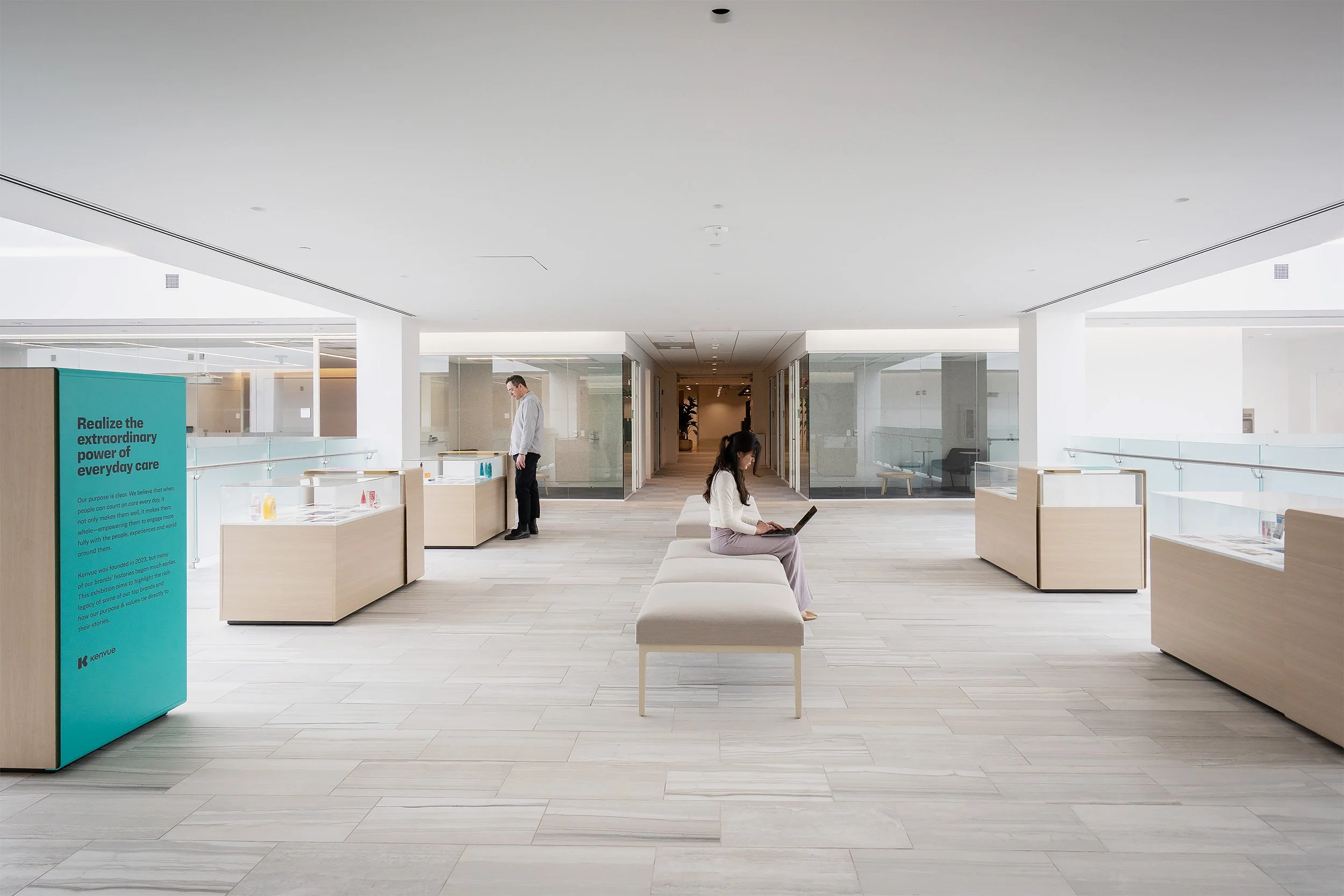 Modern, minimalist office or waiting area with white walls and light wood flooring. There are a few people, one man standing, and a woman sitting on a padded bench working on a laptop. Several display counters and a teal informational sign are visible, along with glass doors leading to different rooms.