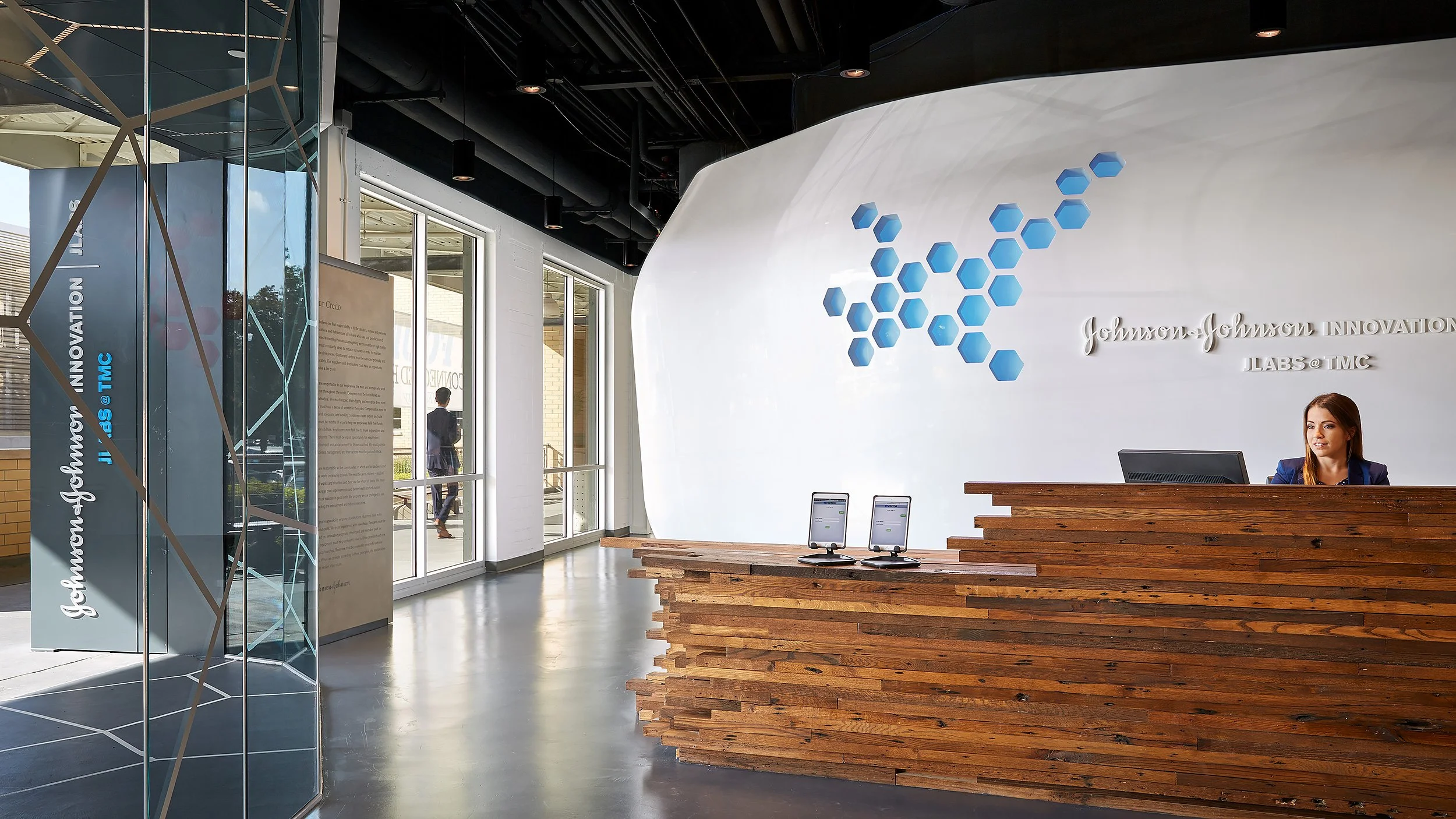 Modern reception area with a wooden front desk, a woman sitting behind a computer, a floral and honeycomb logo on the wall, and glass entrance doors allowing natural light to fill the space.