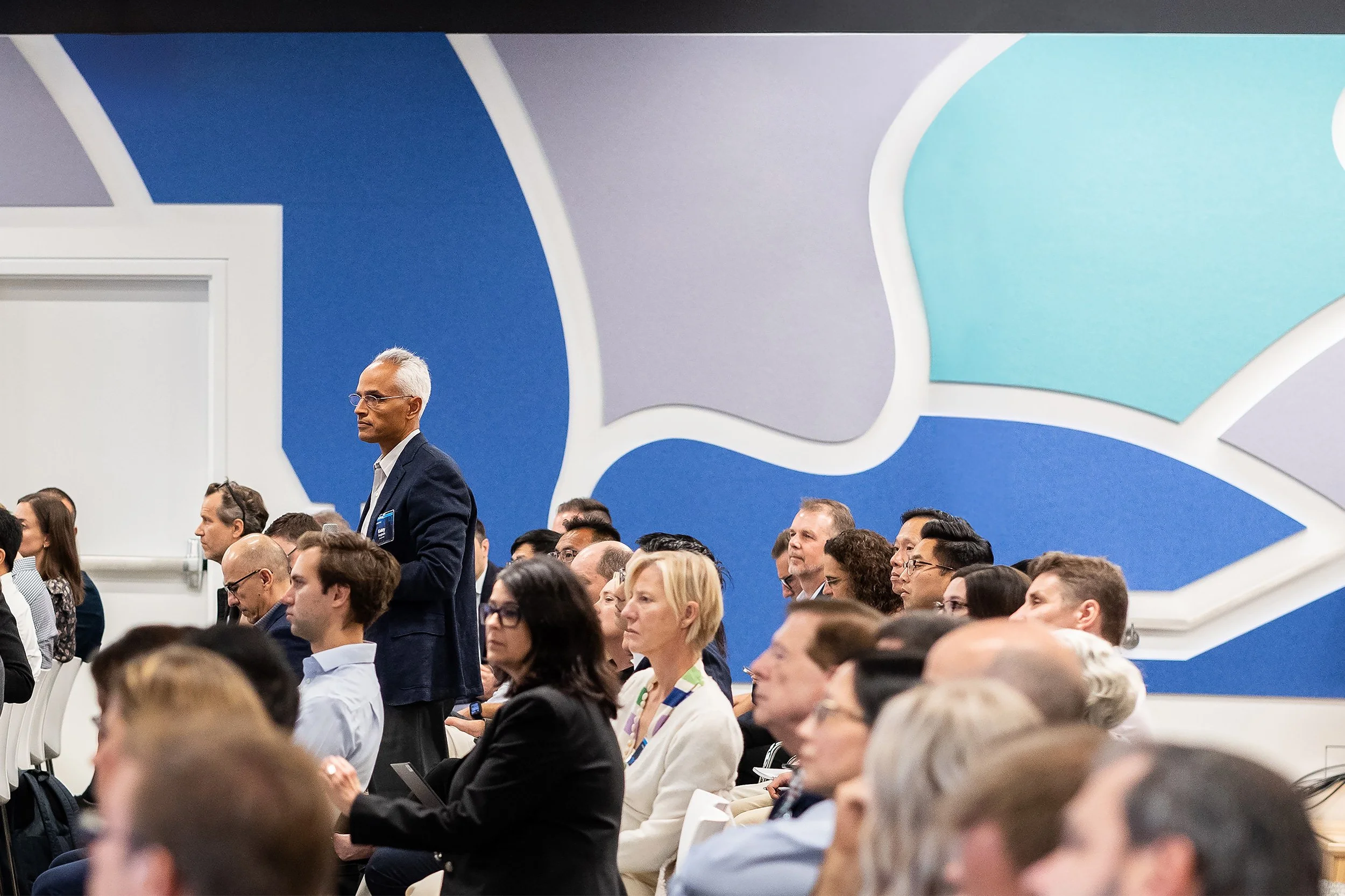 People attending a conference or seminar, sitting in chairs and listening attentively, with a large abstract mural on the wall behind them.