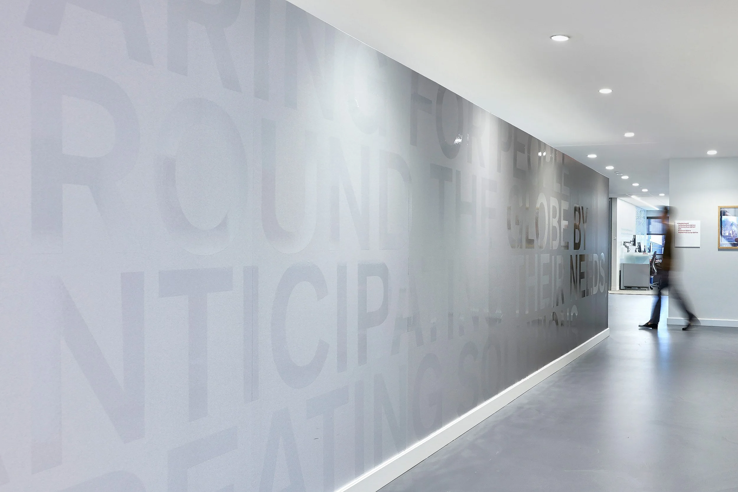 Modern office corridor with a grey wall featuring faint, large text and bright ceiling lights. Two blurred people are visible near the end of the hall.