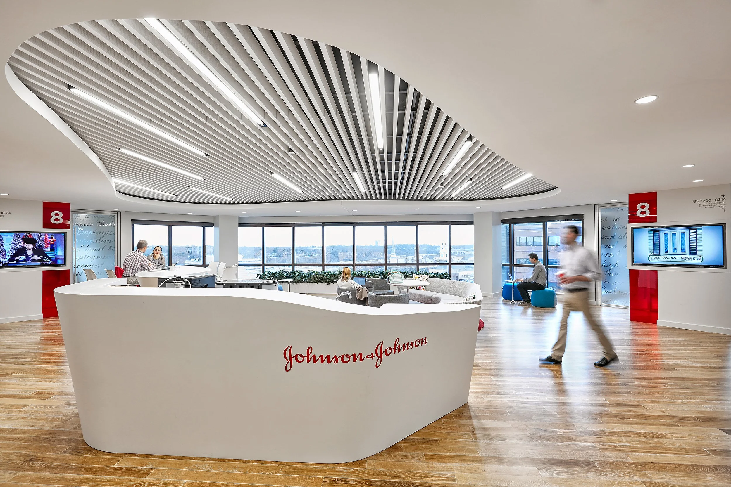 Interior of a modern Johnson & Johnson office with a curved reception desk, multiple seating areas, large windows, and ceiling with linear lighting fixtures.