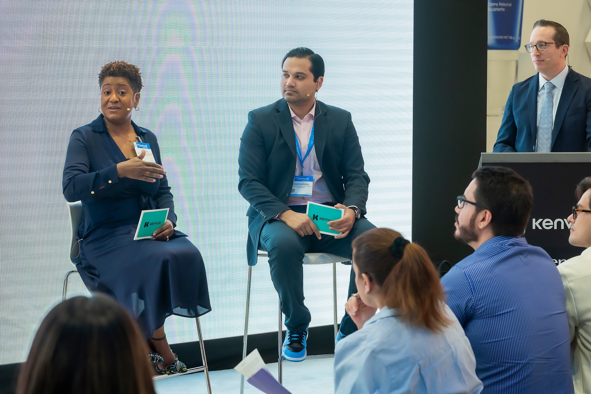 Three panelists, two men and one woman, seated on chairs during a panel discussion at a conference, with an audience listening attentively.