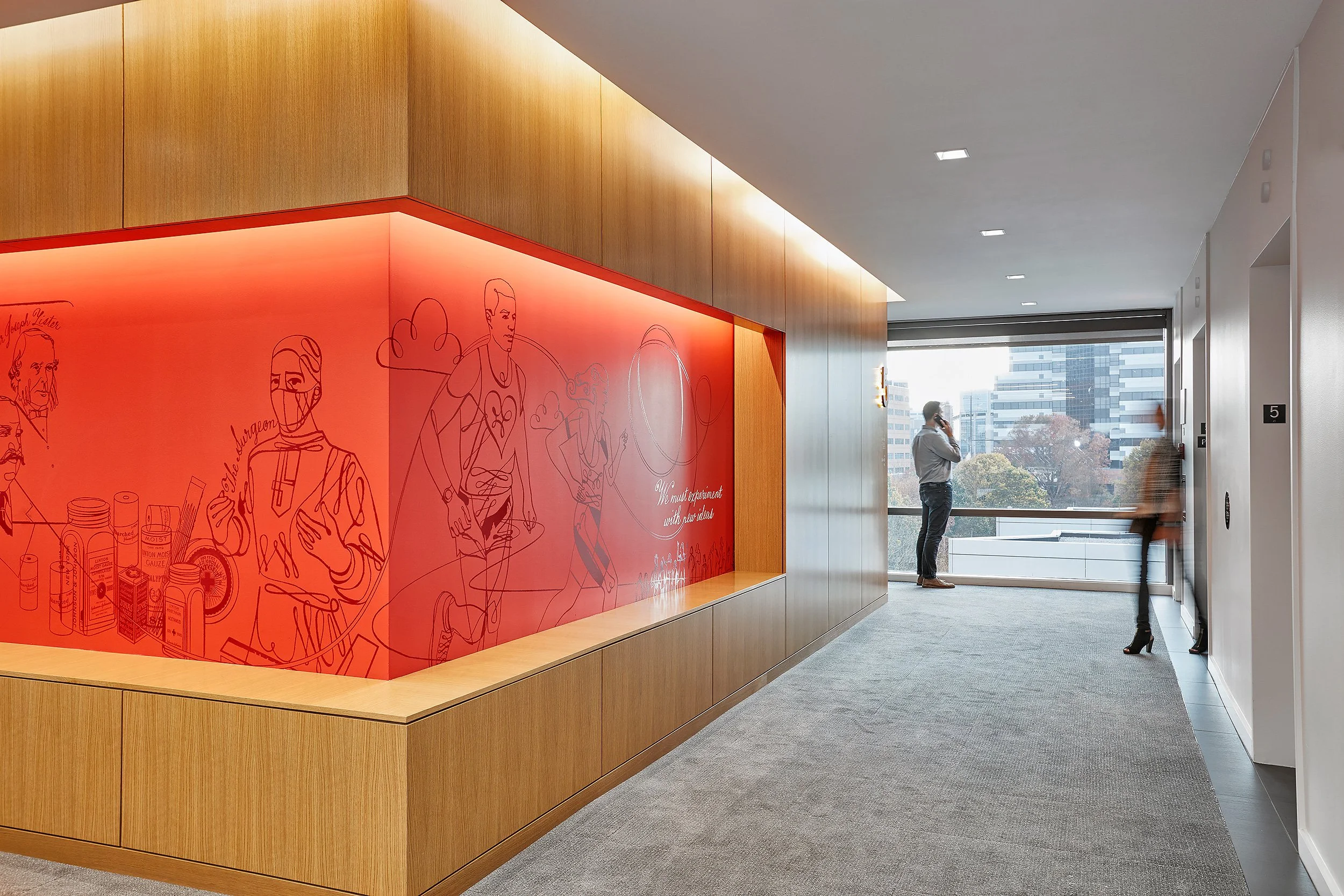 Modern office lobby with wooden wall panels, red wall with illustrations, large window with cityscape view, two people, one talking on phone, one walking past.