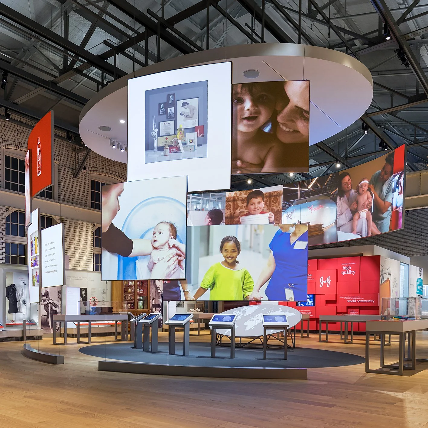 An indoor exhibition space with large digital screens displaying images of children and healthcare scenes. The area features informational kiosks, a red display with the word "high quality", and a globe graphic, suggesting a health or community-themed exhibit.