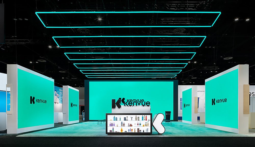 Trade show booth for Kenvue featuring large teal screens, a central display with product samples, and modern lighting design.