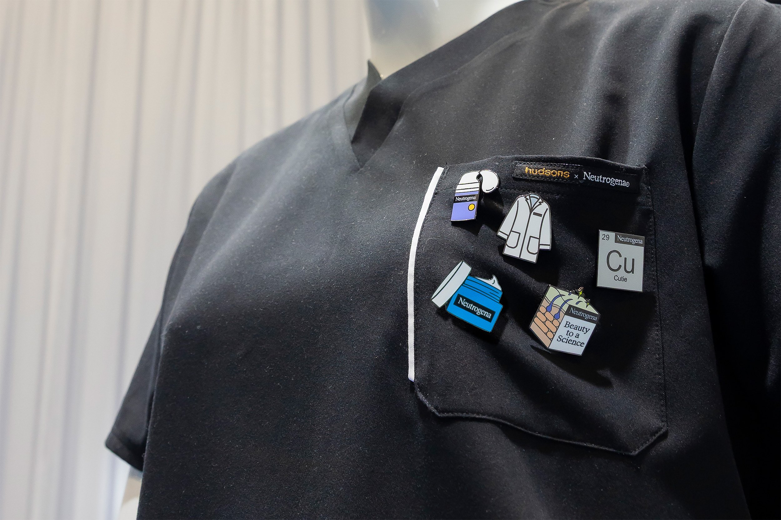 Close-up of a person wearing a black shirt with pins and badges related to Neutrogena skincare products and science, including a doctor illustration and a skincare jar, on the chest pocket.