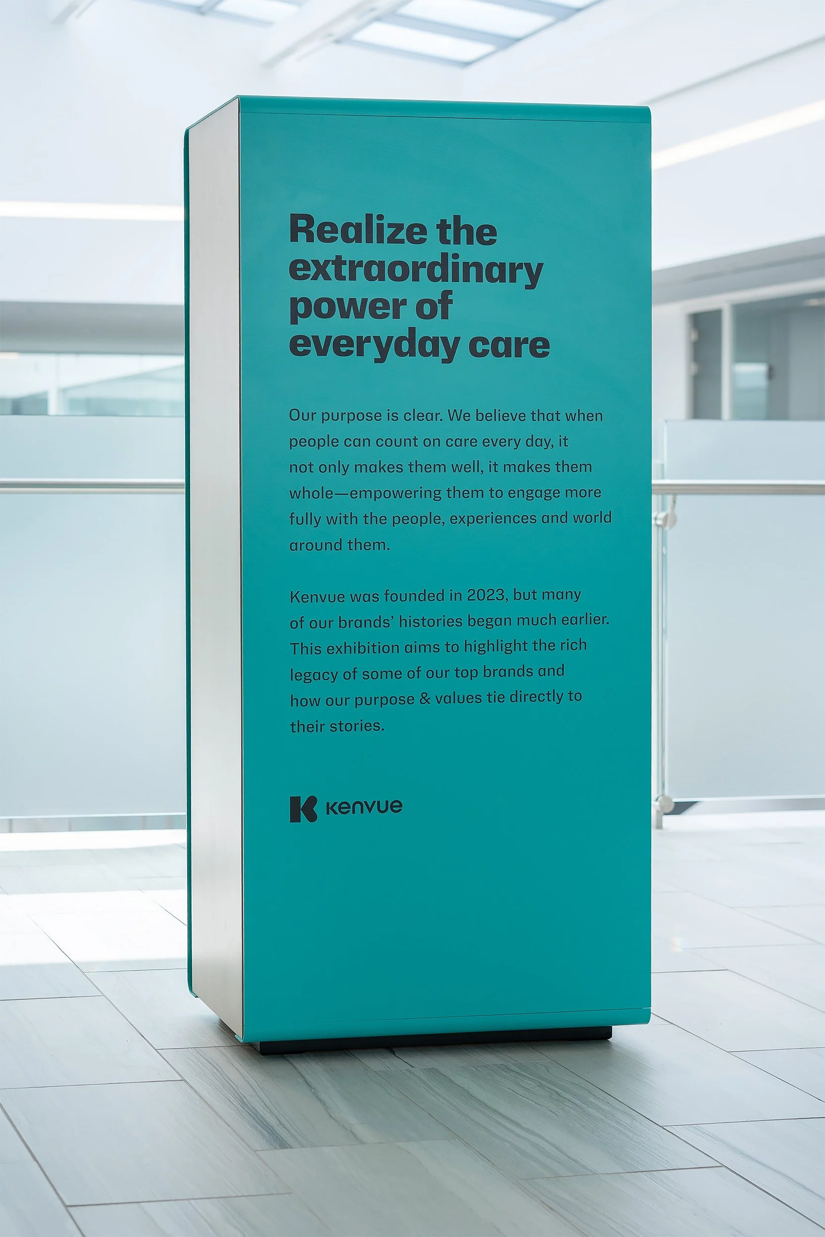 A teal-colored display or sign with the title "Realize the extraordinary power of everyday care" and paragraphs of text underneath. The sign features the Kenveu logo at the bottom, placed in a bright, modern indoor setting with white walls and tiled flooring.