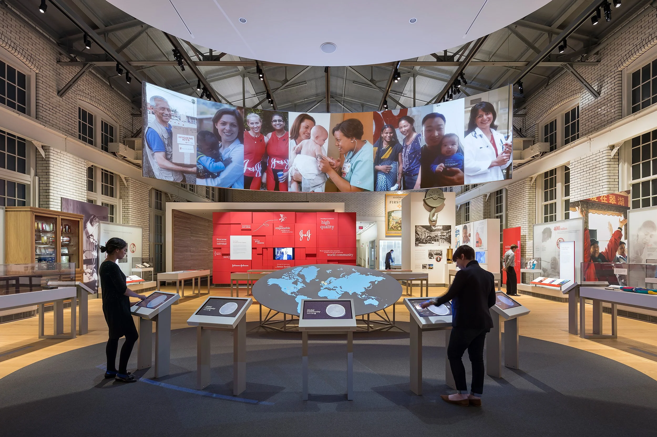 An exhibit space with a large curved display of photographs of diverse people, including children and healthcare workers, above a red informational wall. Visitors are observing displays and a table with a world map at the center.