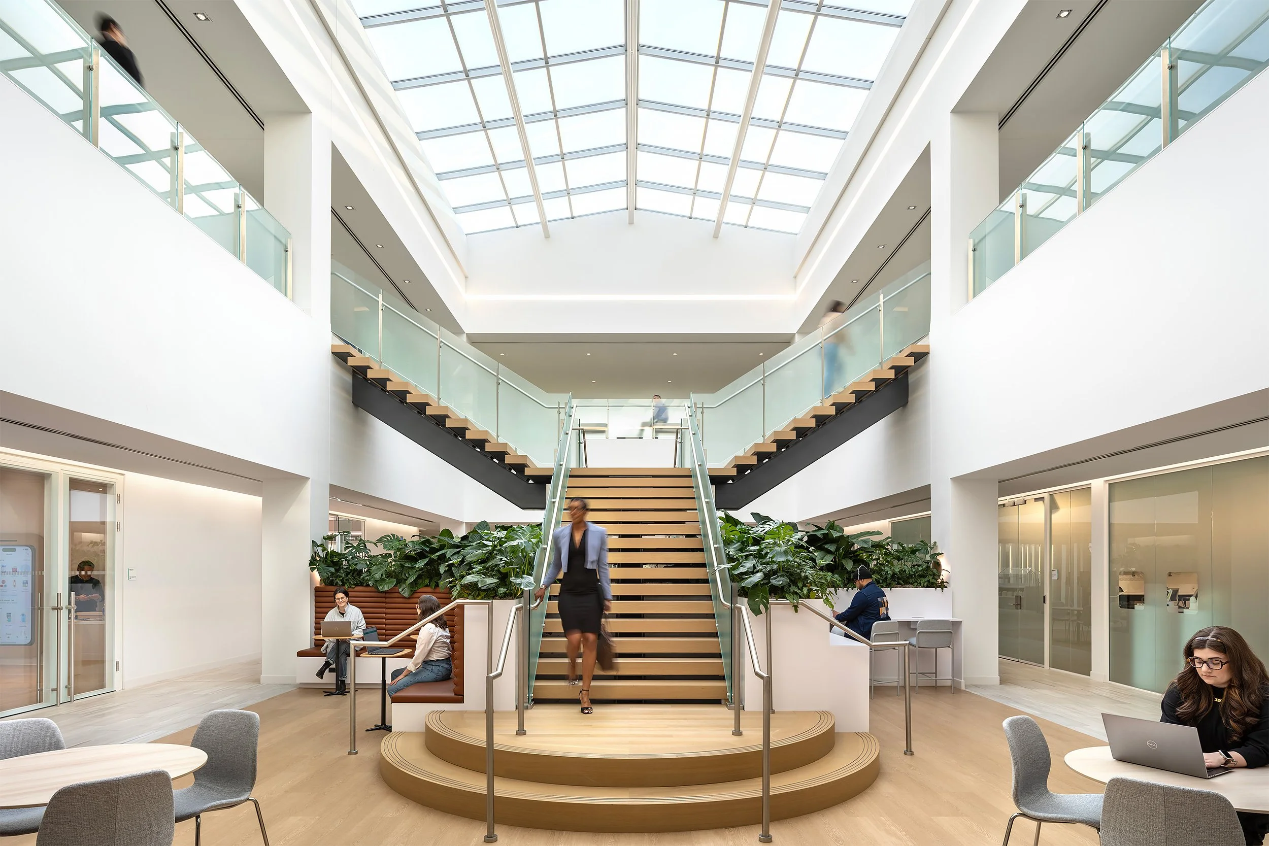 Interior of a modern office building with a central staircase, glass railings, and large skylight ceiling, people working and walking around.