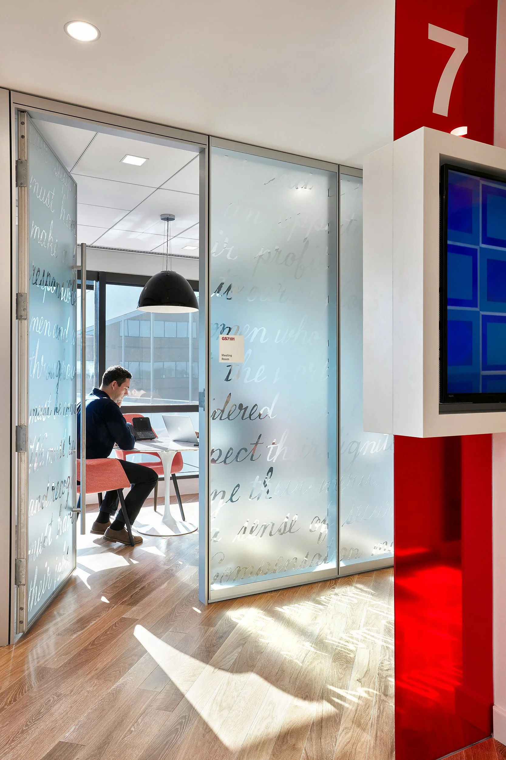 An office meeting room with a man sitting at a round table, working on a laptop, inside a glass-walled room with handwritten script on the glass. The room is near a bright red pillar displaying the number 7. Sunlight streams onto the woodenfloor.