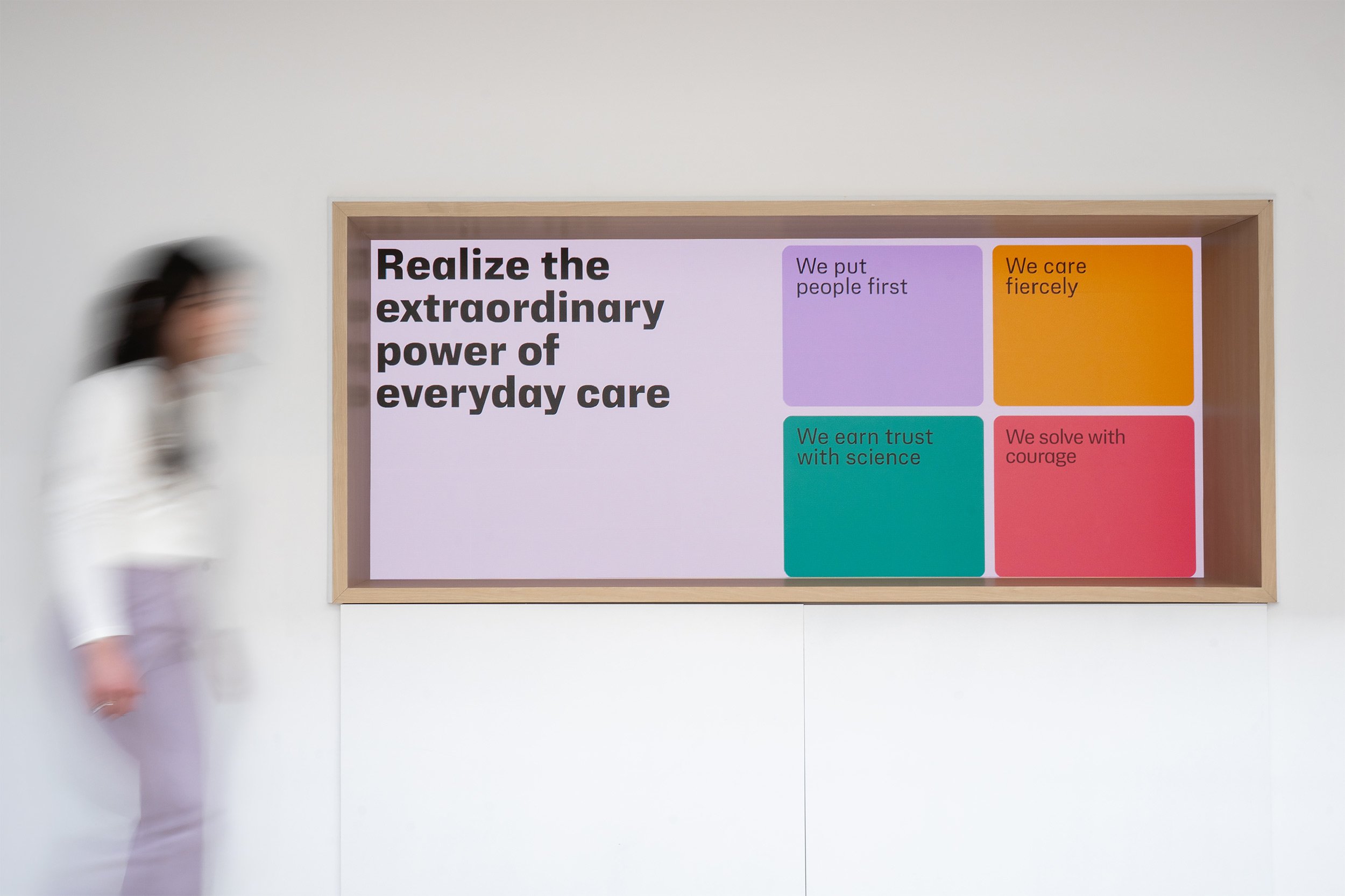 Colorful informational display on a white wall featuring statements about everyday care, trust, science, and courage, with a blurred person walking past.