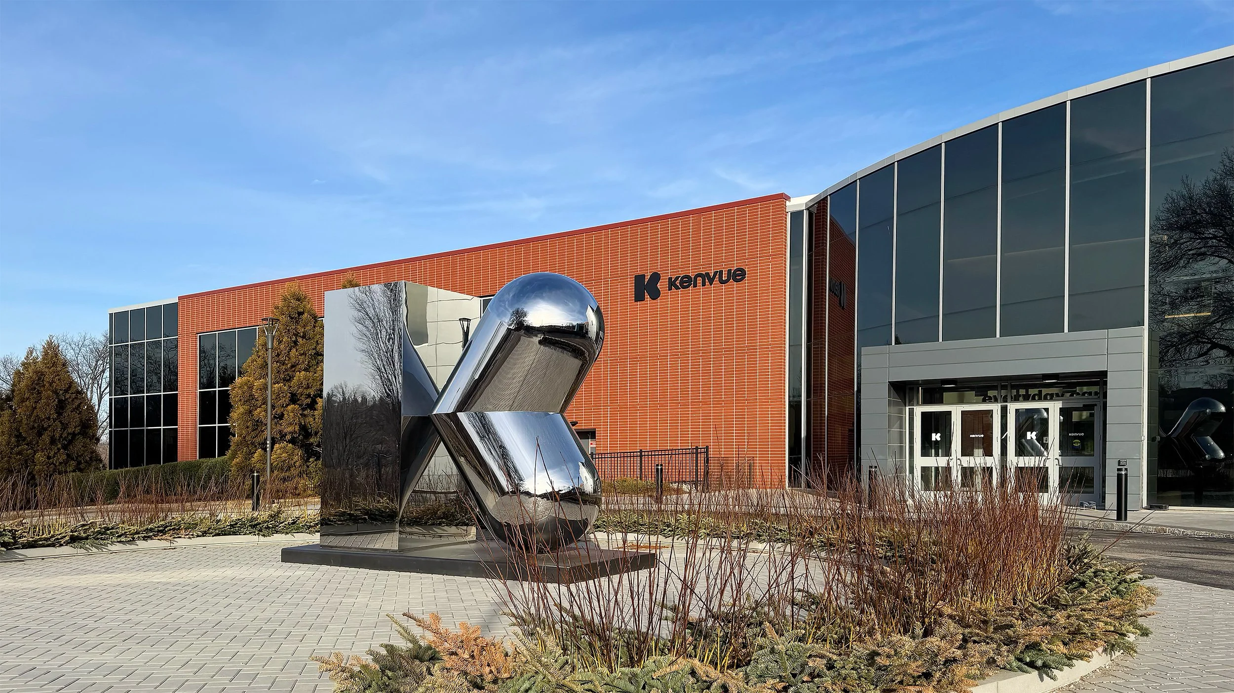 Modern building with red brick and glass windows, the Kenvue logo, and a reflective metallic sculpture in front.