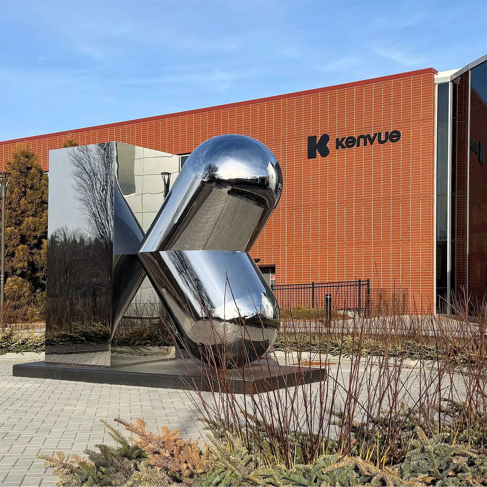 Modern building with red brick exterior and large signage reading 'Kenvue'. In front, an abstract metallic sculpture with reflective surface and rounded shapes. Landscaping with dried plants and trees, clear blue sky.
