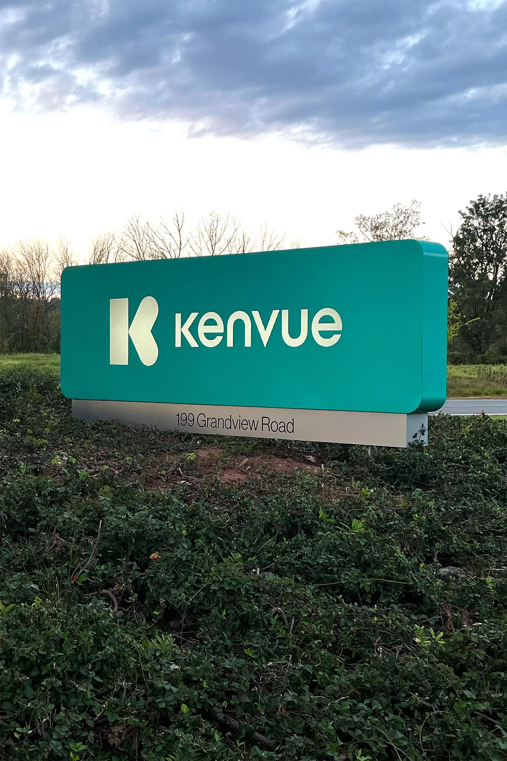 A teal-colored Kenvue sign with white text and logo, located on a grassy area with bushes in front, and trees and a cloudy sky in the background, at 199 Grandview Road.