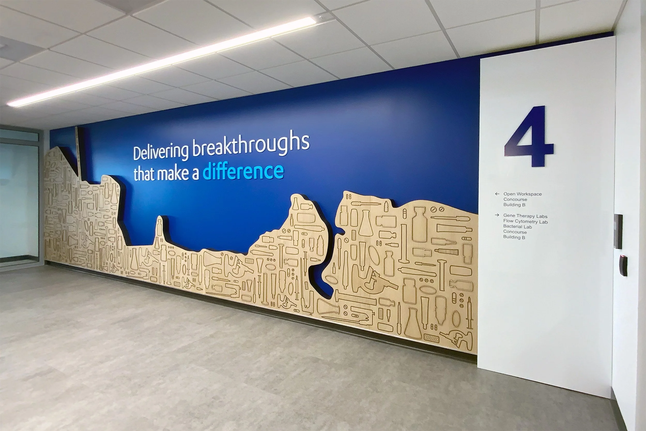 Interior view of a hallway with a large mural on the wall. The mural has a blue background with white text that reads 'Delivering breakthroughs that make a difference' and a large purple number 4. There are wooden shapes with black outlines resembling laboratory tools and scientific equipment on the mural.